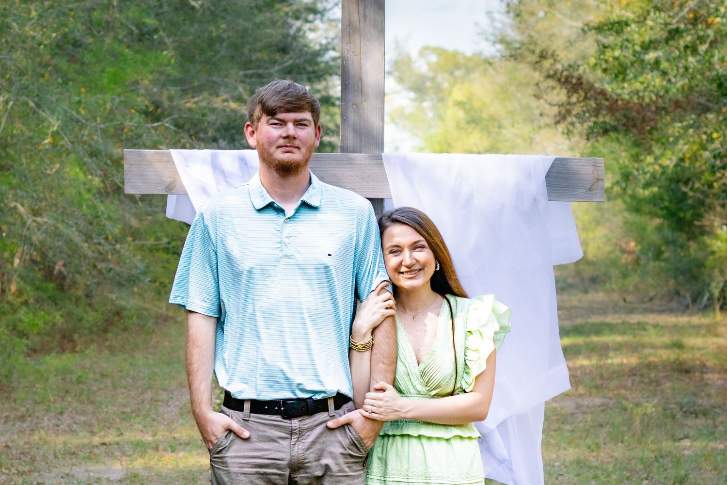 Young couple standing before a wooden cross outdoors, man in blue polo, woman in green dress leaning on him.