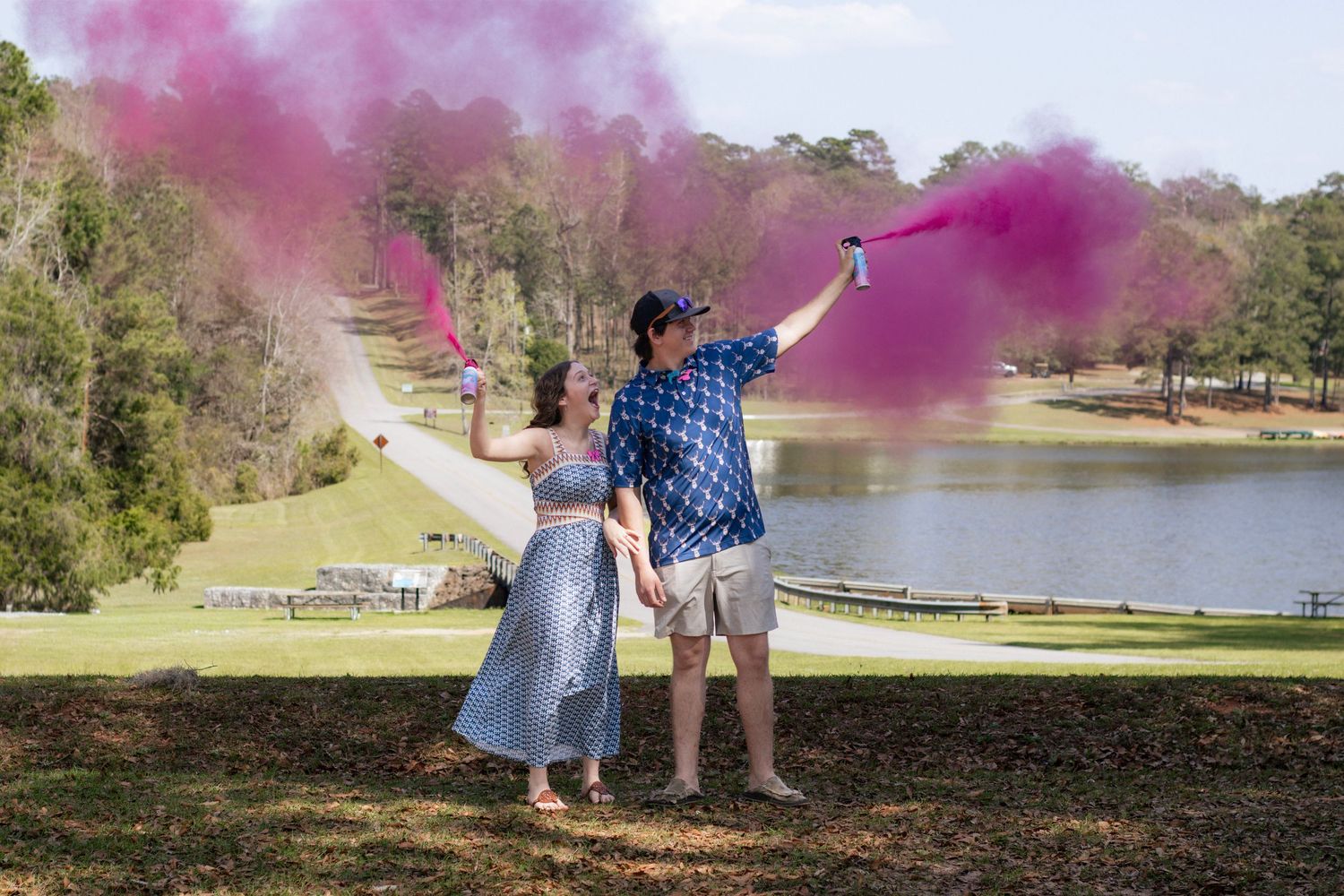 Couple holds pink smoke cannons during outdoor gender reveal by a lake.