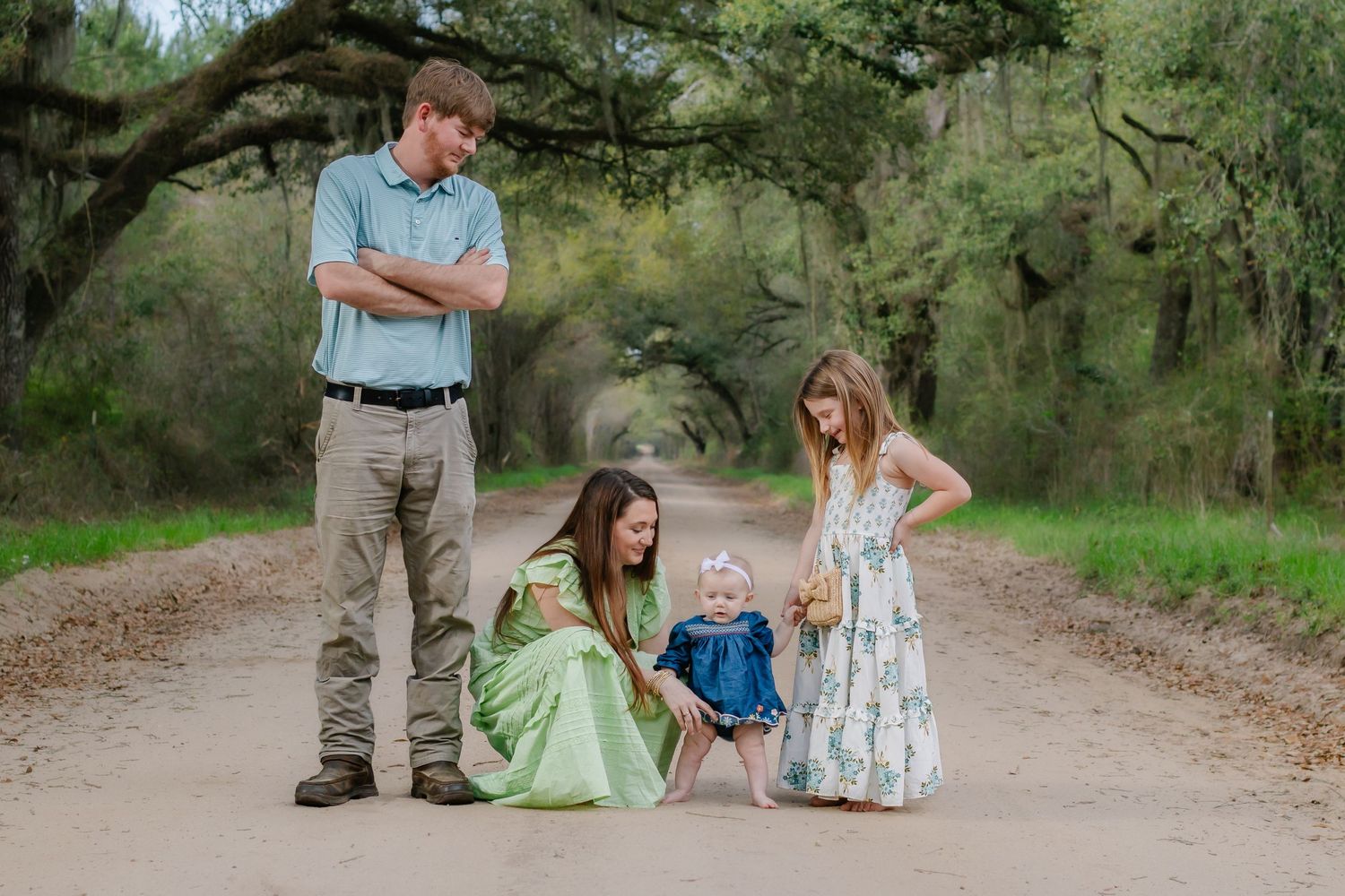 Family with young children posing on a rustic dirt road under a natural canopy of green trees.