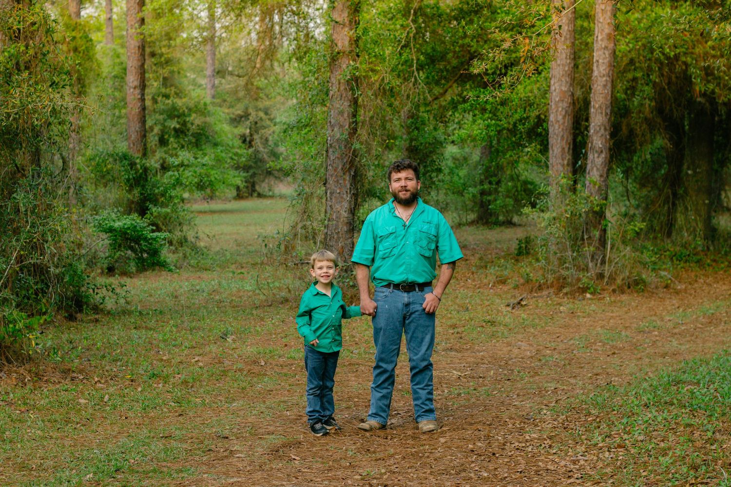 A father and young son in matching green shirts hold hands while standing on a forest path.