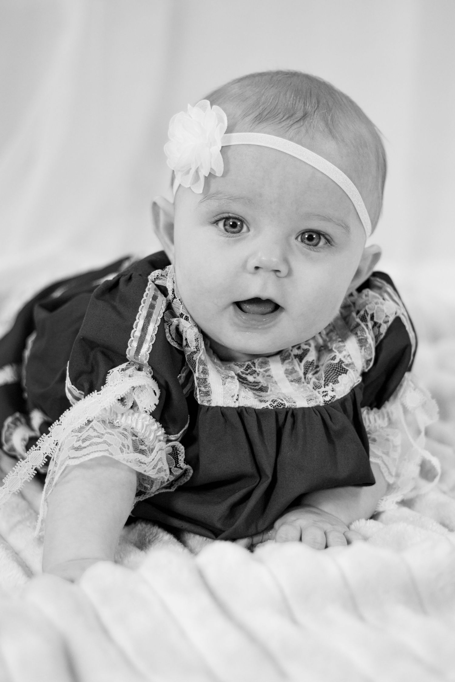 Black and white portrait of a baby girl in a lace dress and white floral headband lying on a blanket.