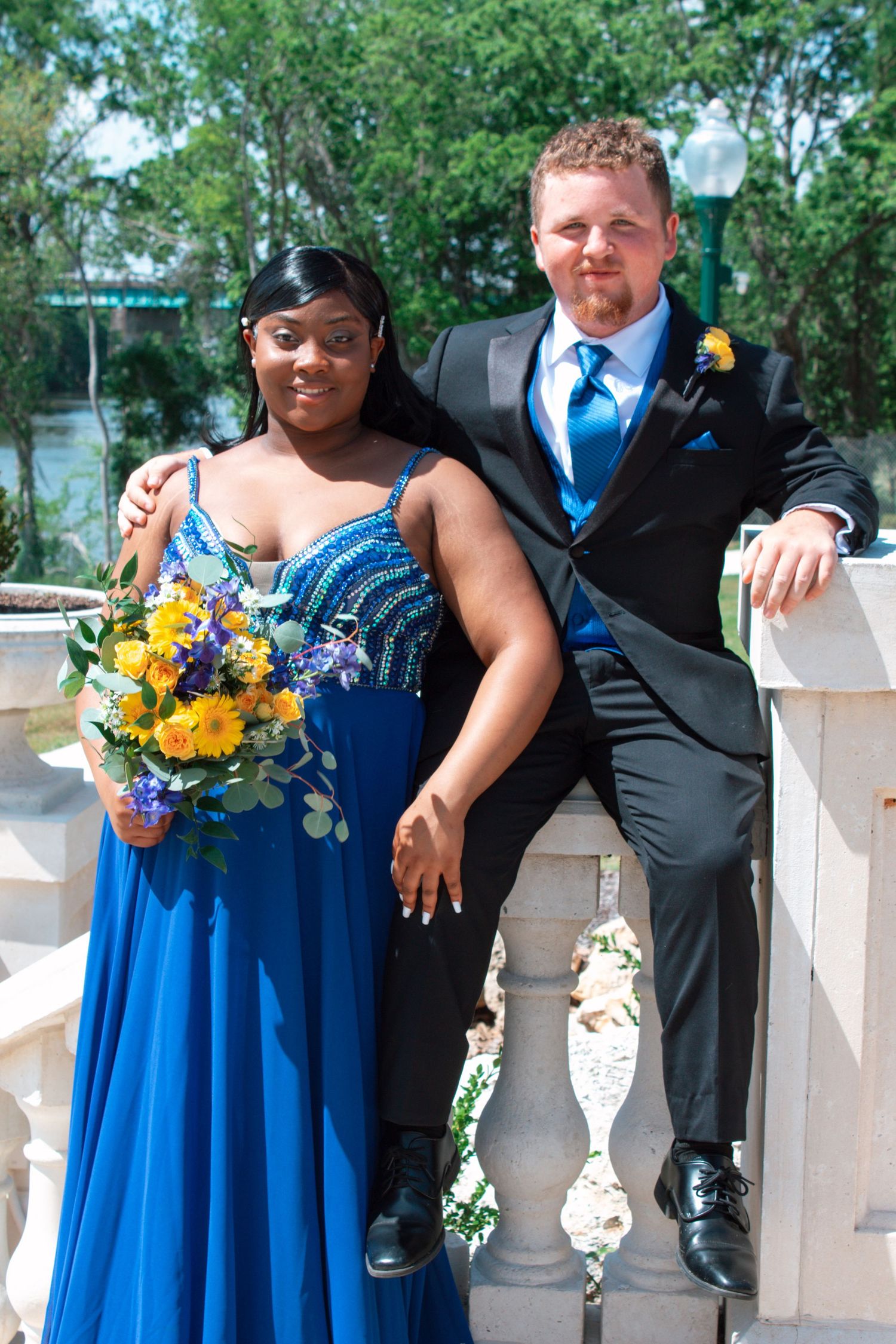 Couple in formal prom attire posing outdoors, woman in blue gown holding flowers, man in black suit.