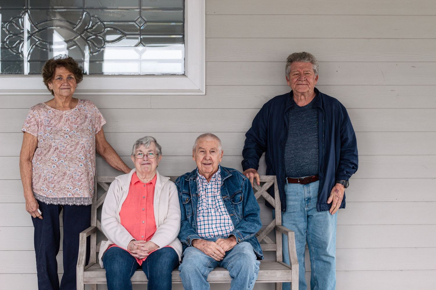 Four seniors pose together on a porch, two standing and two seated on a wooden bench.