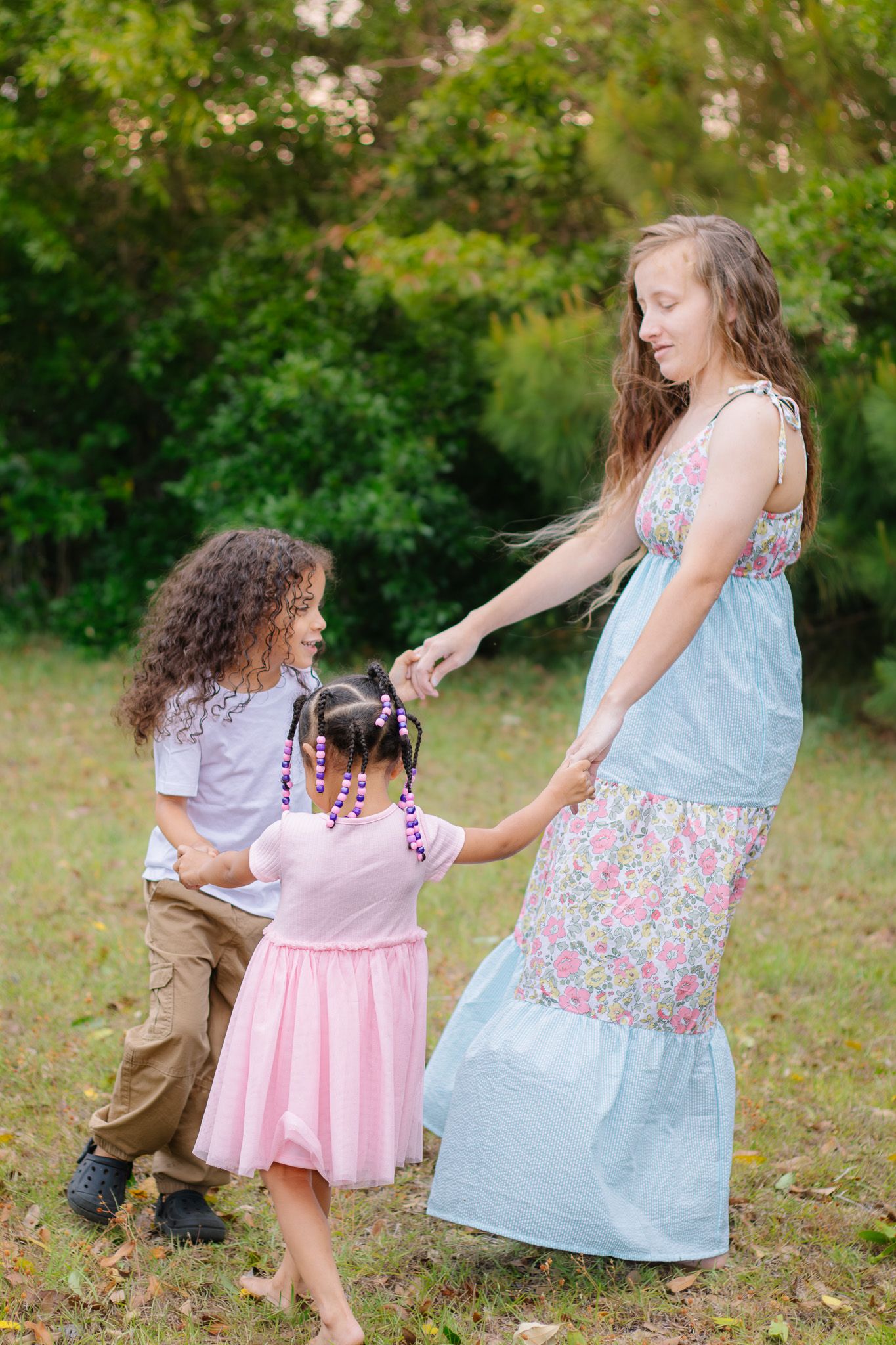 A teenage girl in a floral dress plays with two young children outdoors in a green park setting.