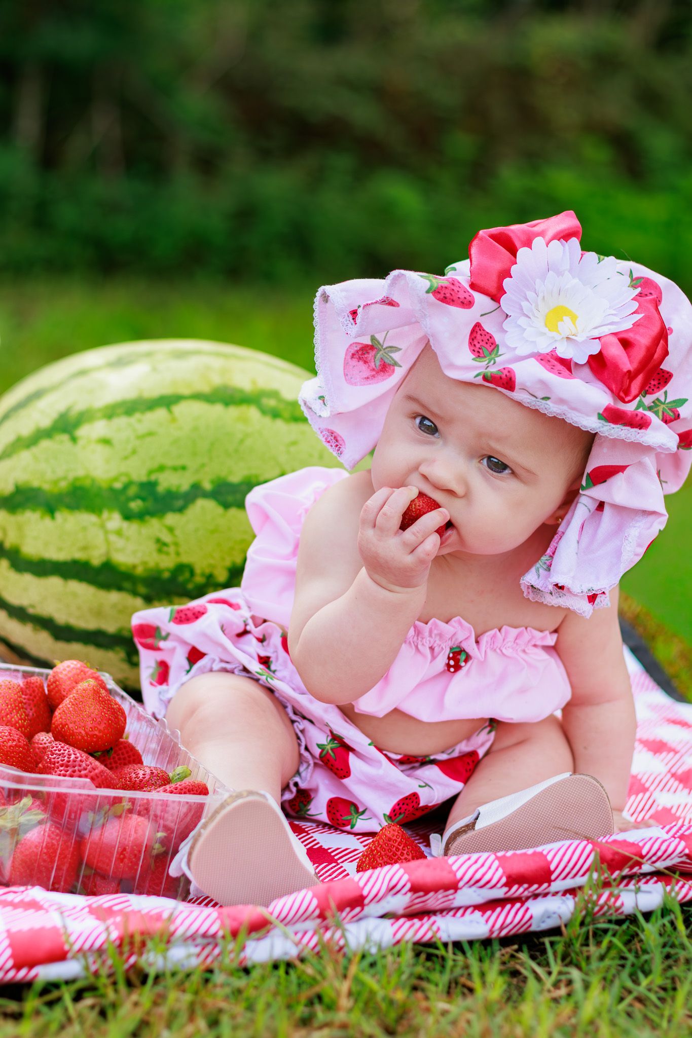 Cute baby in pink strawberry hat and matching outfit eating strawberries on a red checkered blanket outdoors.