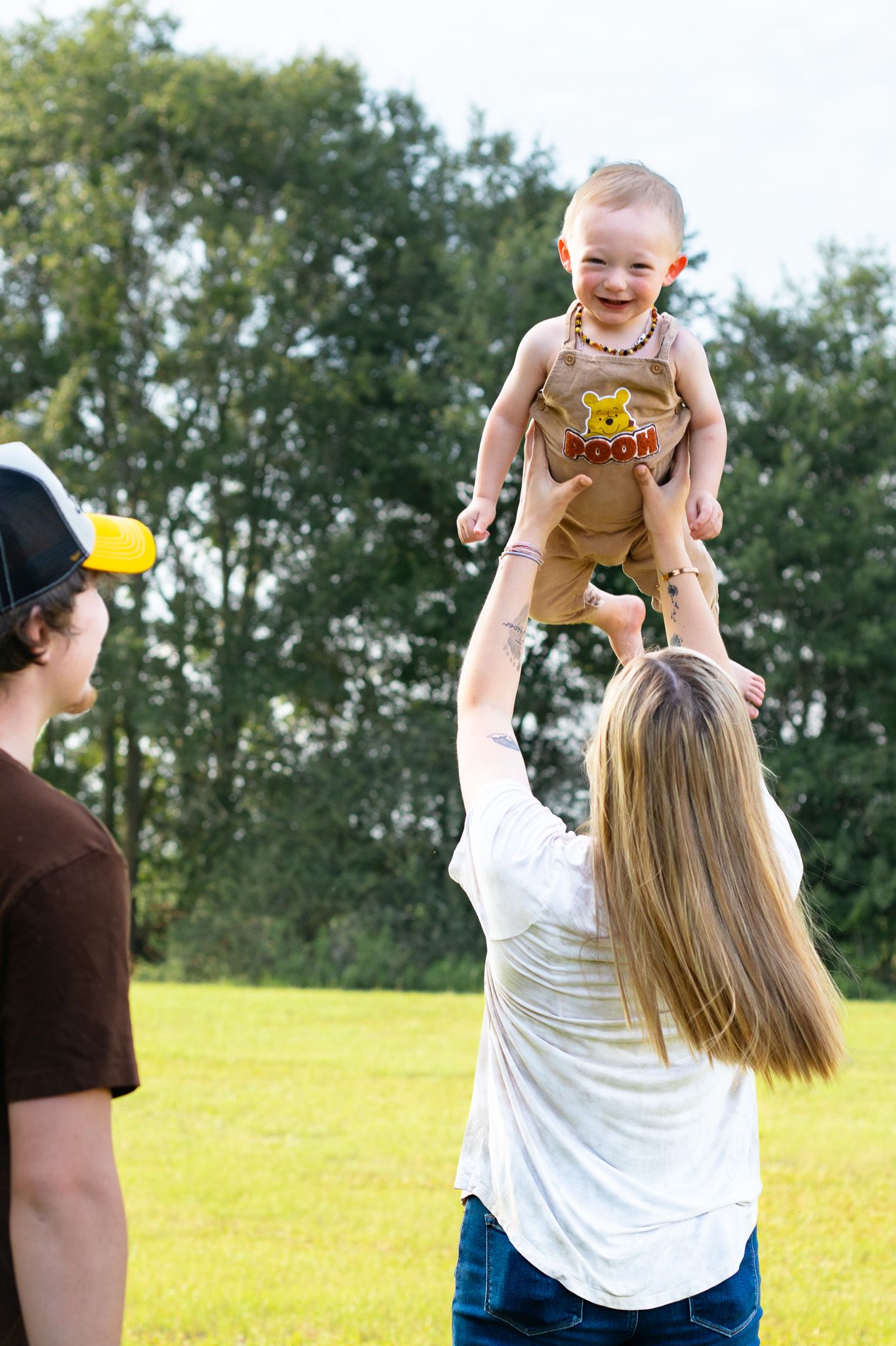 A joyful mother lifts her laughing baby high in the air outdoors while a man watches nearby.