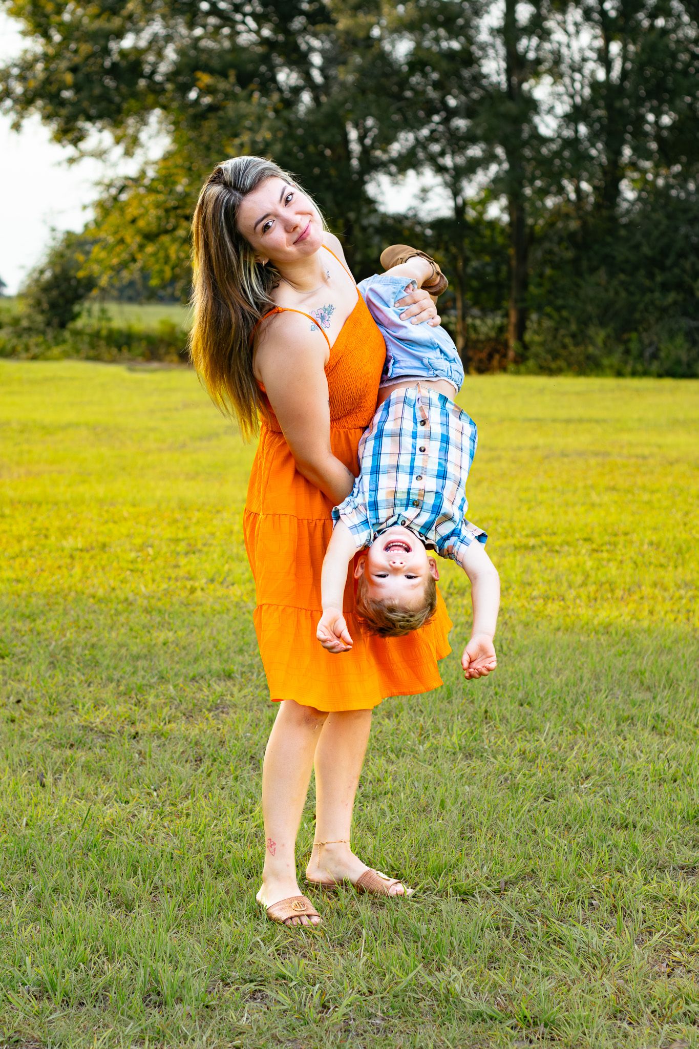 A smiling woman in an orange dress holds a laughing child upside down in a sunny green park.