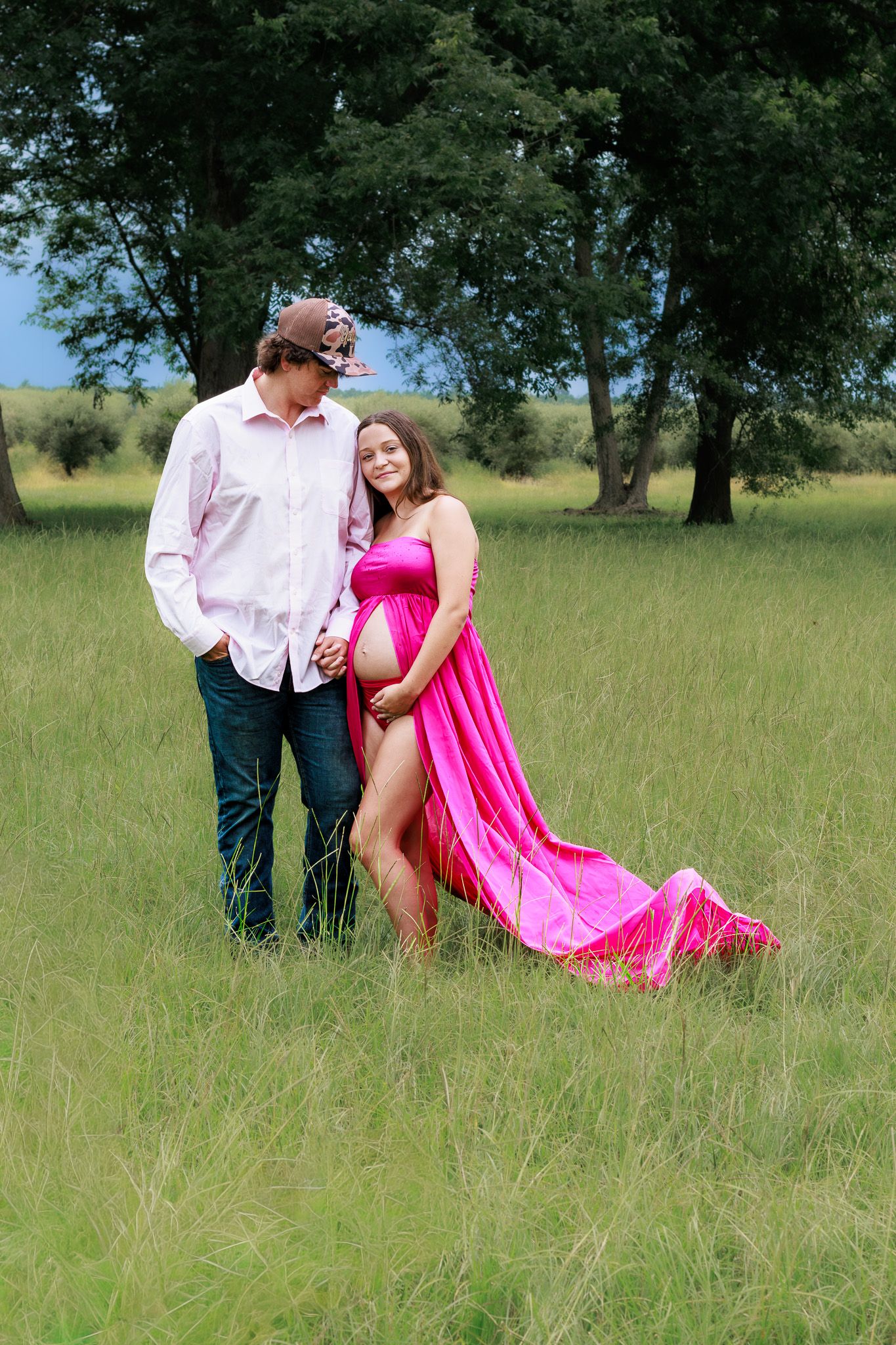 Pregnant woman in pink flowing gown stands with partner in lush green field during outdoor maternity photoshoot.