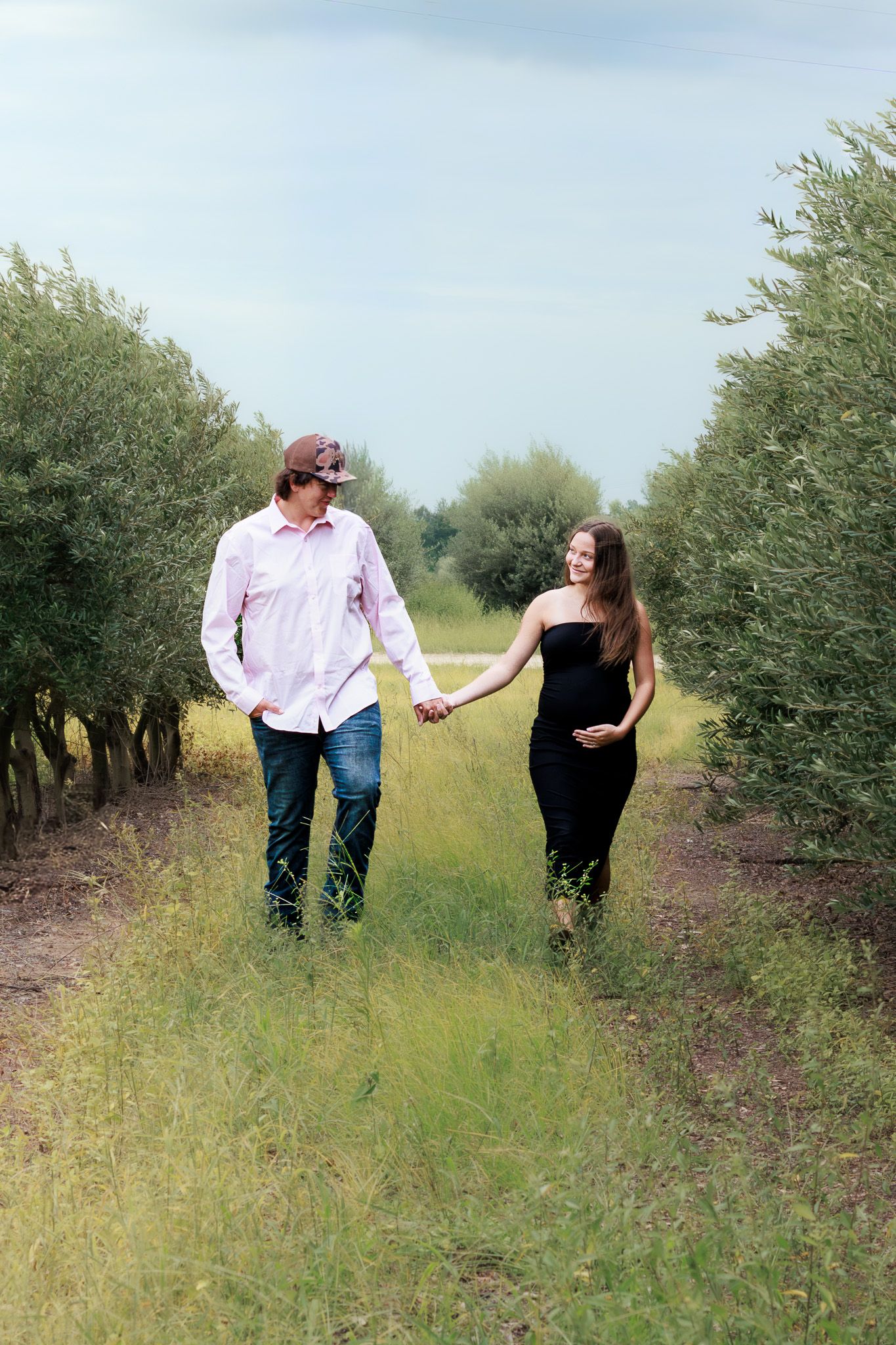 Pregnant couple holding hands and walking through a lush green orchard path, smiling at each other on a cloudy day.