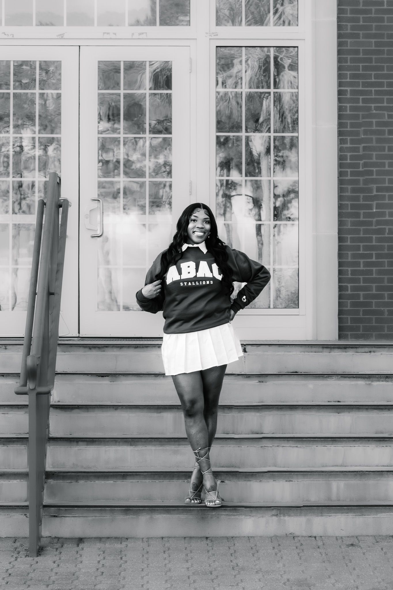 Black and white photo of a smiling young woman in ABAU sweatshirt and white skirt posing on steps.