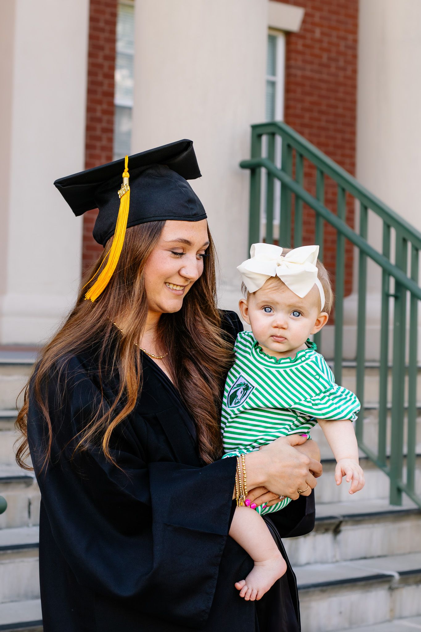 Graduate in cap and gown smiling while holding a baby girl with a white bow headband outdoors.