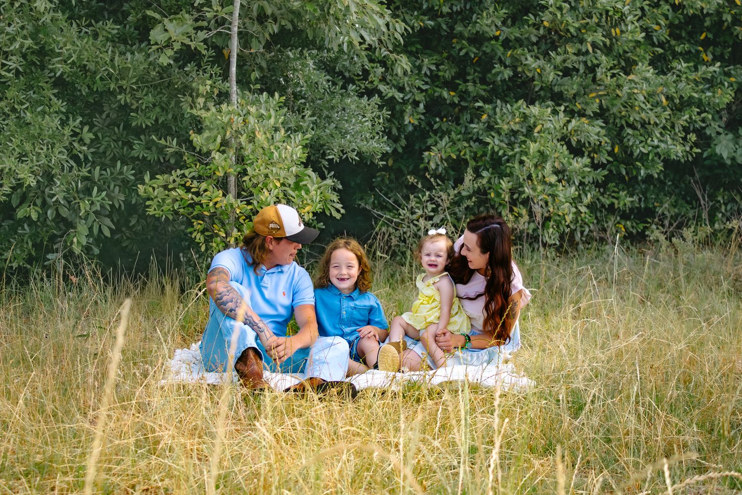 Family of five sitting on a blanket in a sunny meadow surrounded by lush green trees, smiling and enjoying time together.