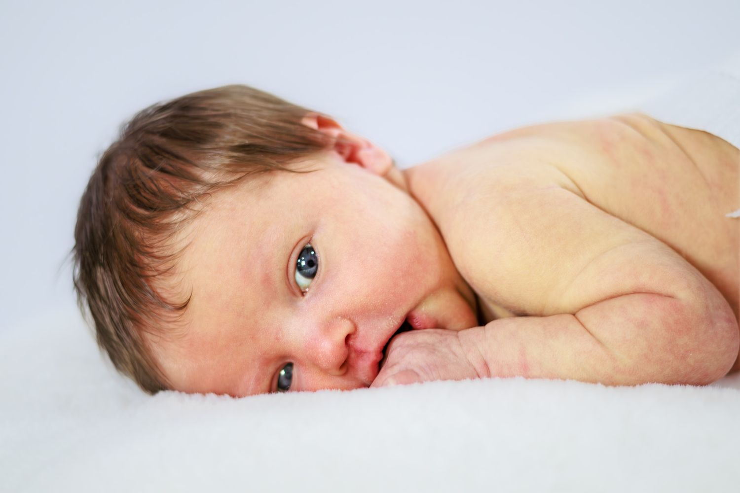Newborn baby with blue eyes lying on white blanket looking at camera.