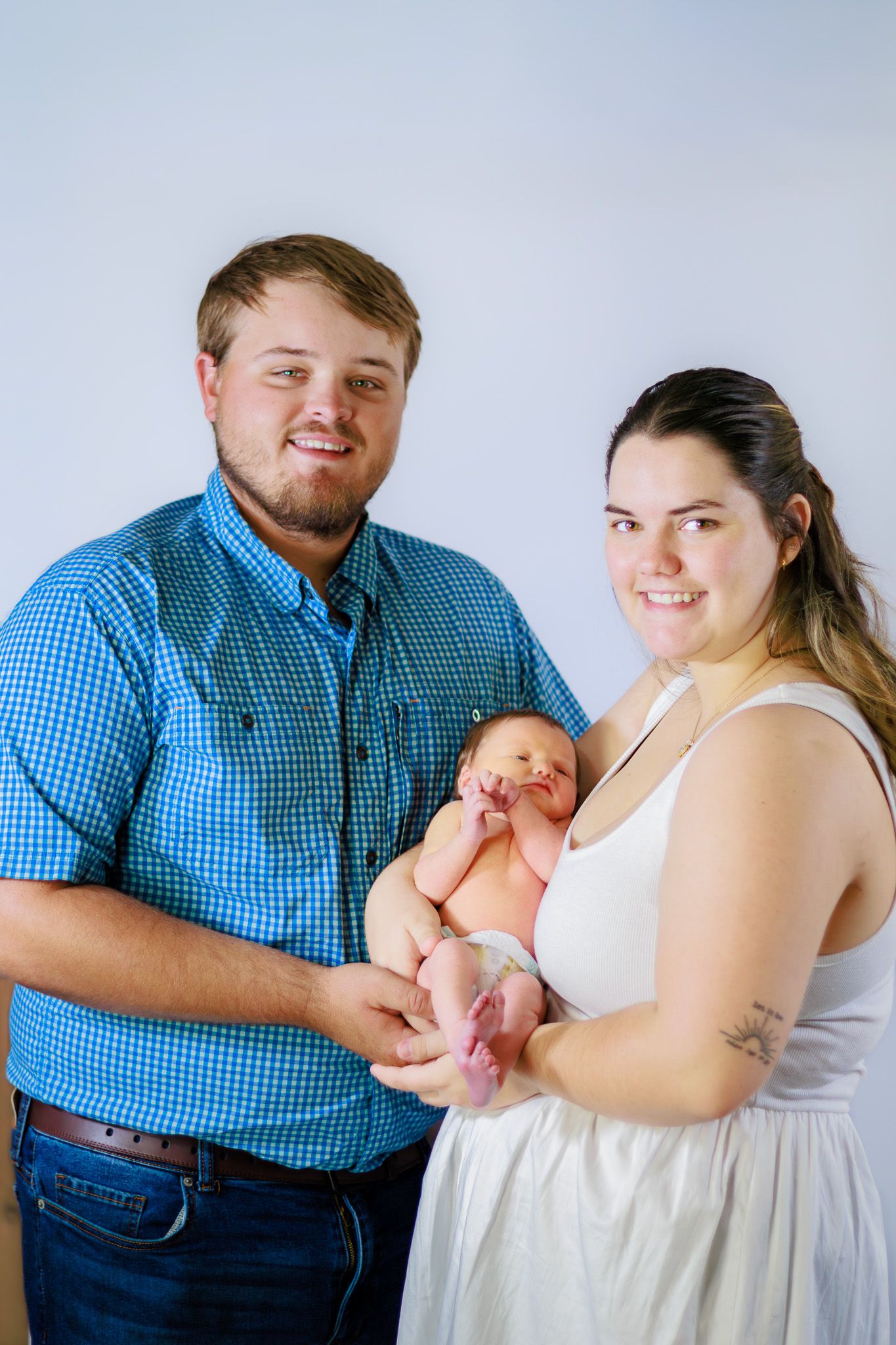 Smiling couple holding their newborn baby, man in blue shirt, woman in white dress.