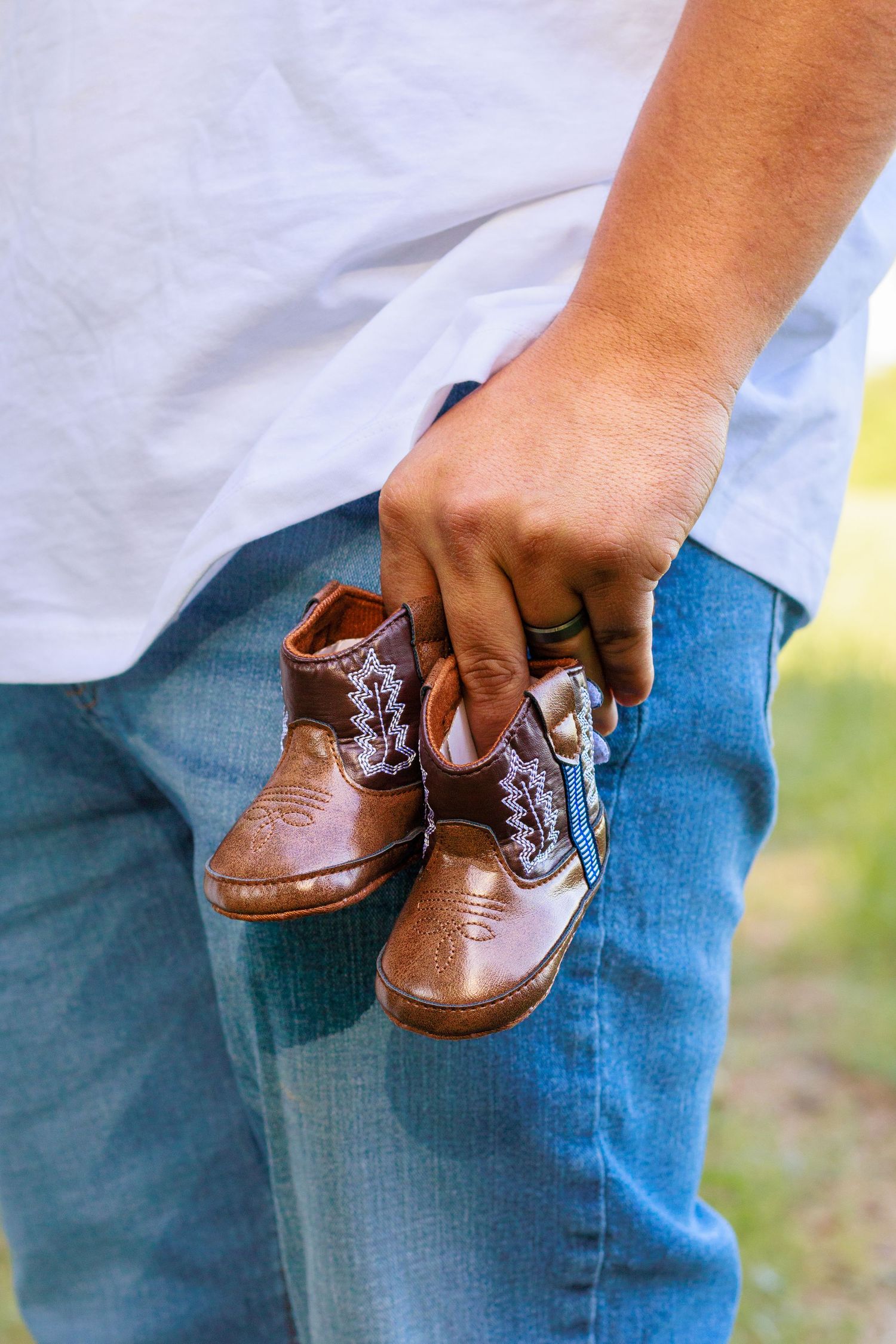 Person holding tiny brown leather baby cowboy boots while wearing jeans and a white t-shirt outdoors.