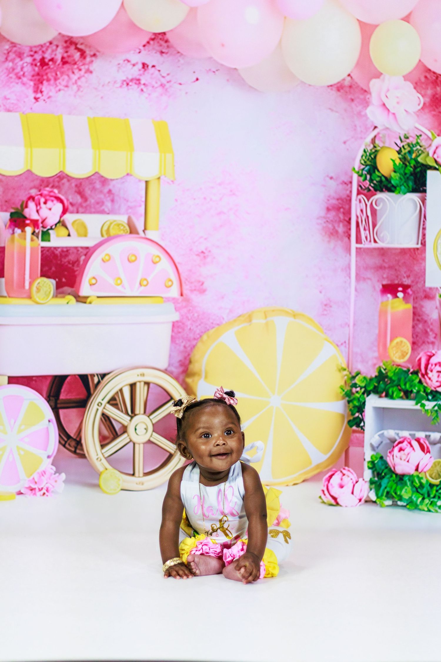 Smiling baby girl in pink outfit sits before a lemonade stand backdrop with balloons and floral decorations.