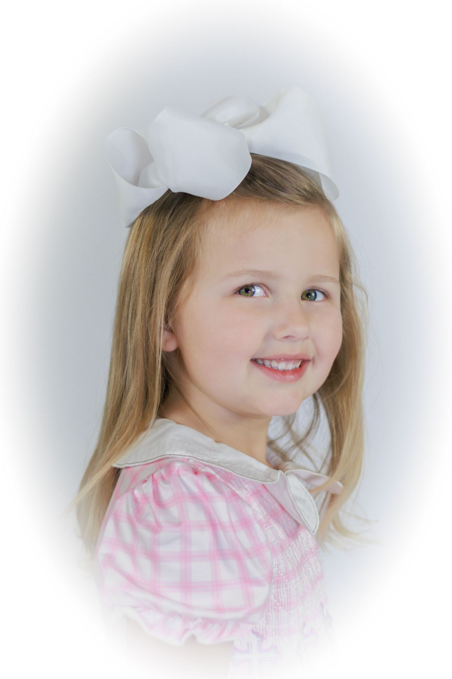 Young girl with blonde hair wearing a white bow and pink gingham smocked dress, smiling over her shoulder.