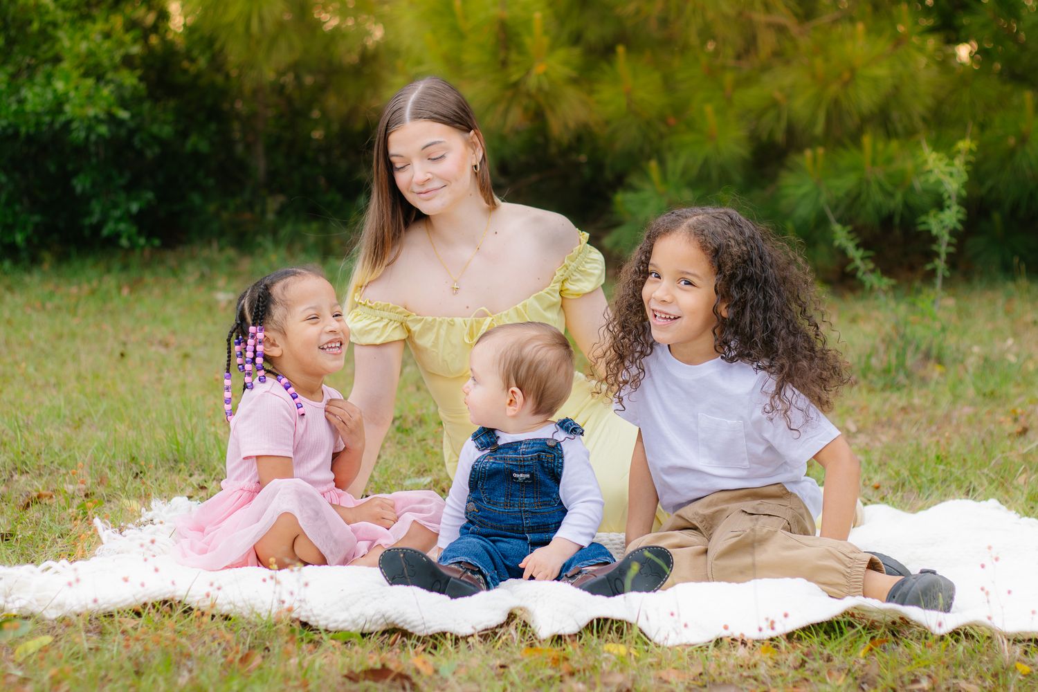A young woman sits on a blanket outdoors with three small children smiling in a grassy park.