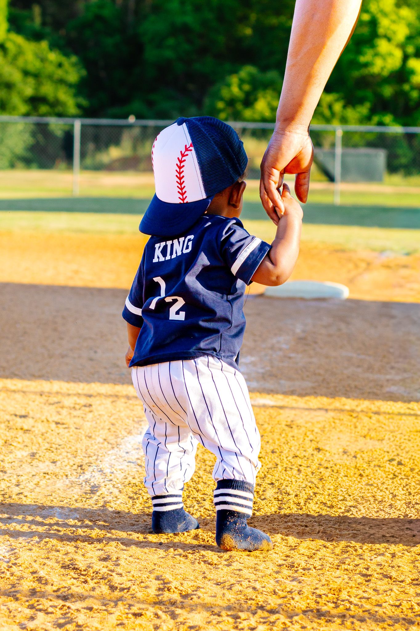 Toddler in King baseball jersey holding adult hand on sunny baseball diamond.