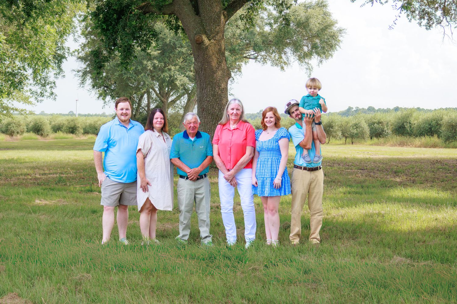 Family of seven posing outdoors near a large tree on a green lawn during a summer portrait session.