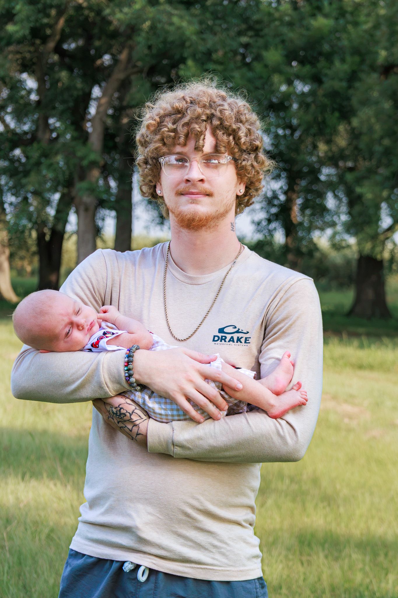 Young curly-haired man with glasses holding a newborn baby outdoors in a green park setting.