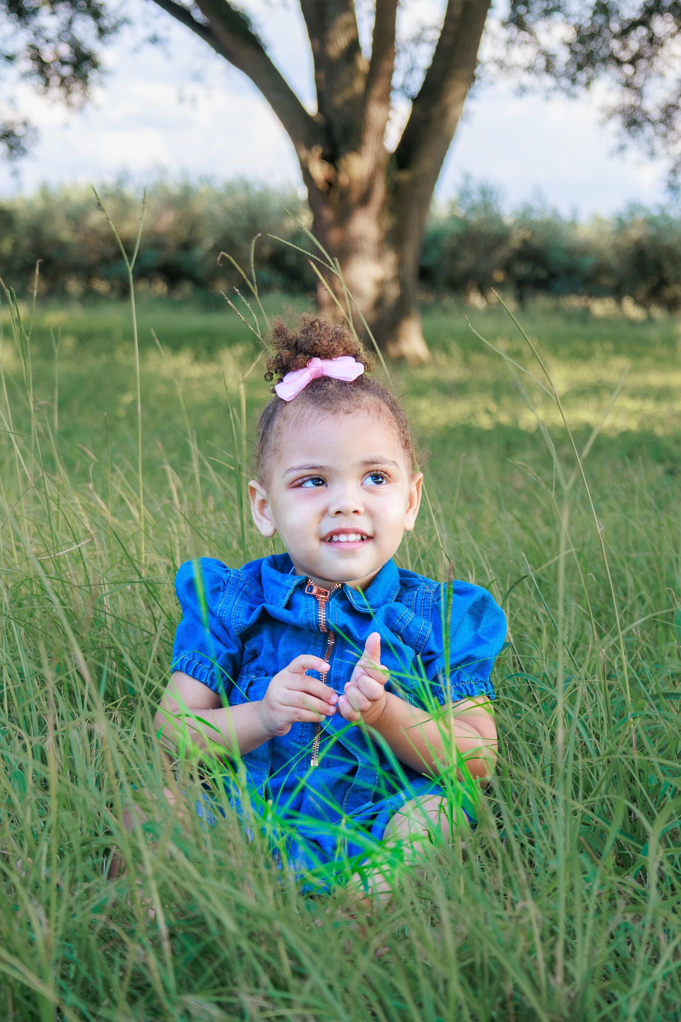 Smiling young girl in blue dress sitting in tall green grass with a pink bow in her curly hair.
