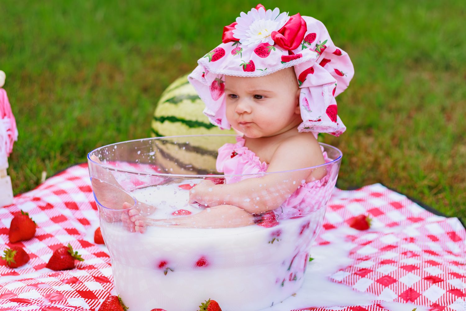 Baby girl in strawberry hat sits in milk bath bowl on red checkered blanket outdoors with strawberries.