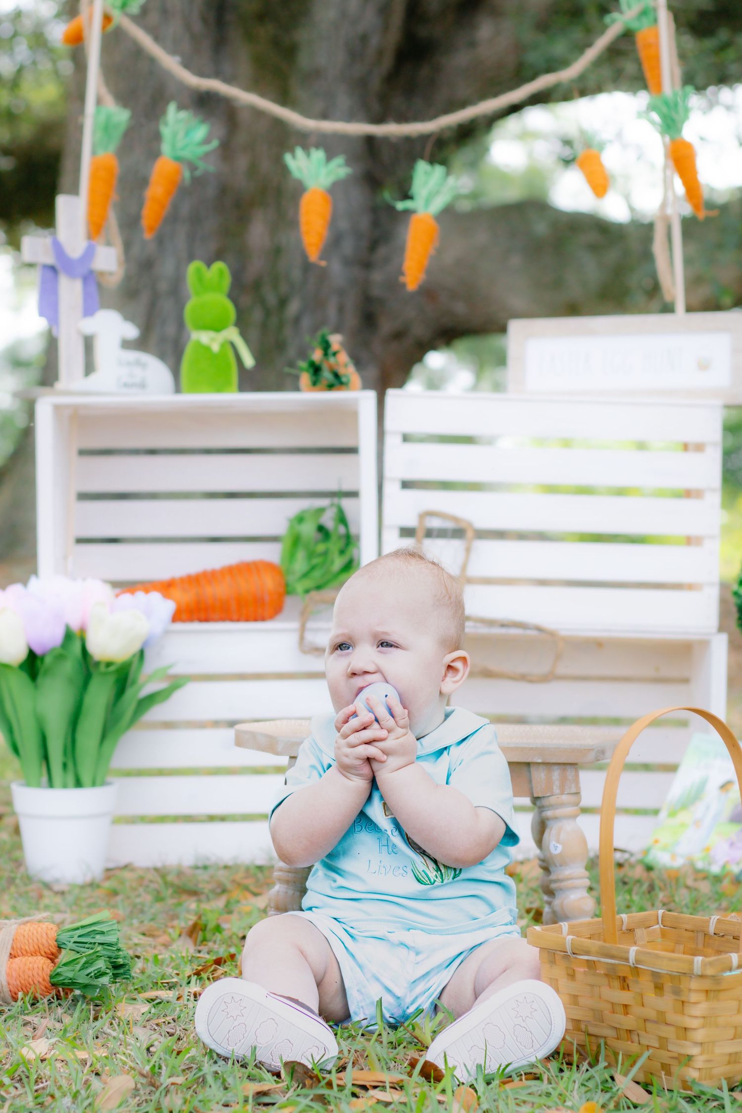 Baby boy in blue romper holds Easter egg while sitting near basket and spring decorations.