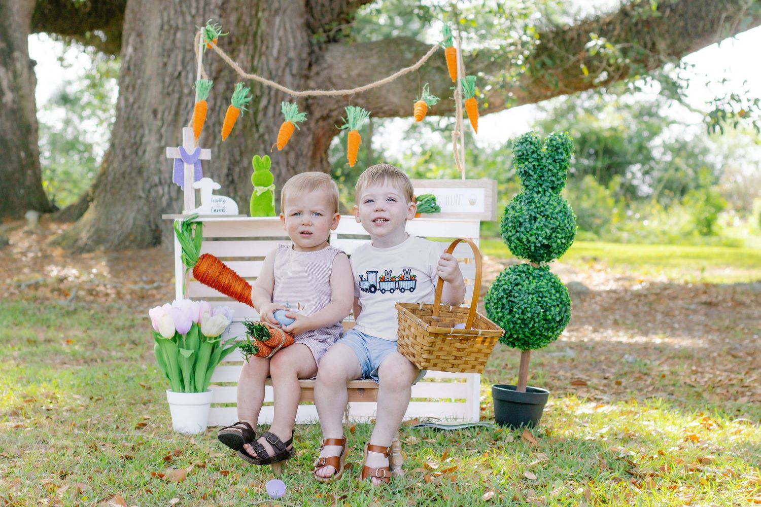 Two toddlers sitting on white bench with Easter carrot garland and topiary tree in outdoor setting.