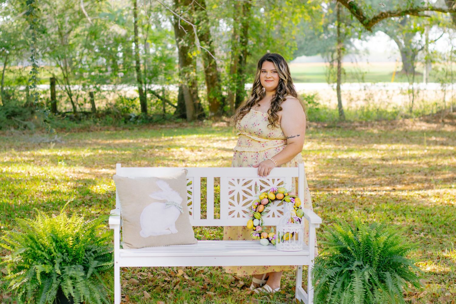 Woman in floral dress poses beside white bench with Easter wreath and ferns in park.