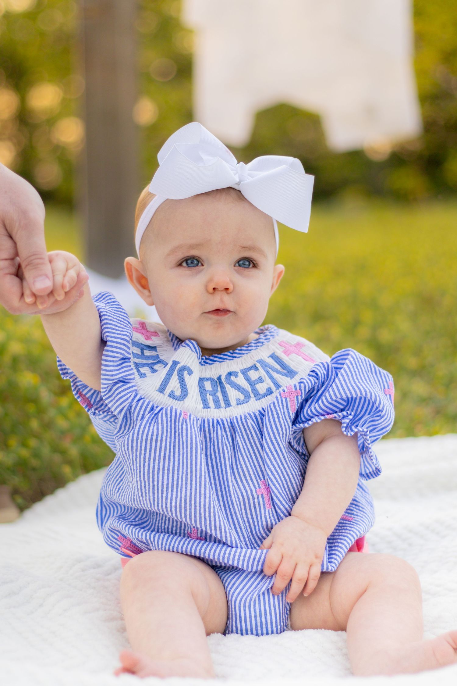Adorable baby in blue striped He Is Risen outfit with white bow headband sitting on white blanket.