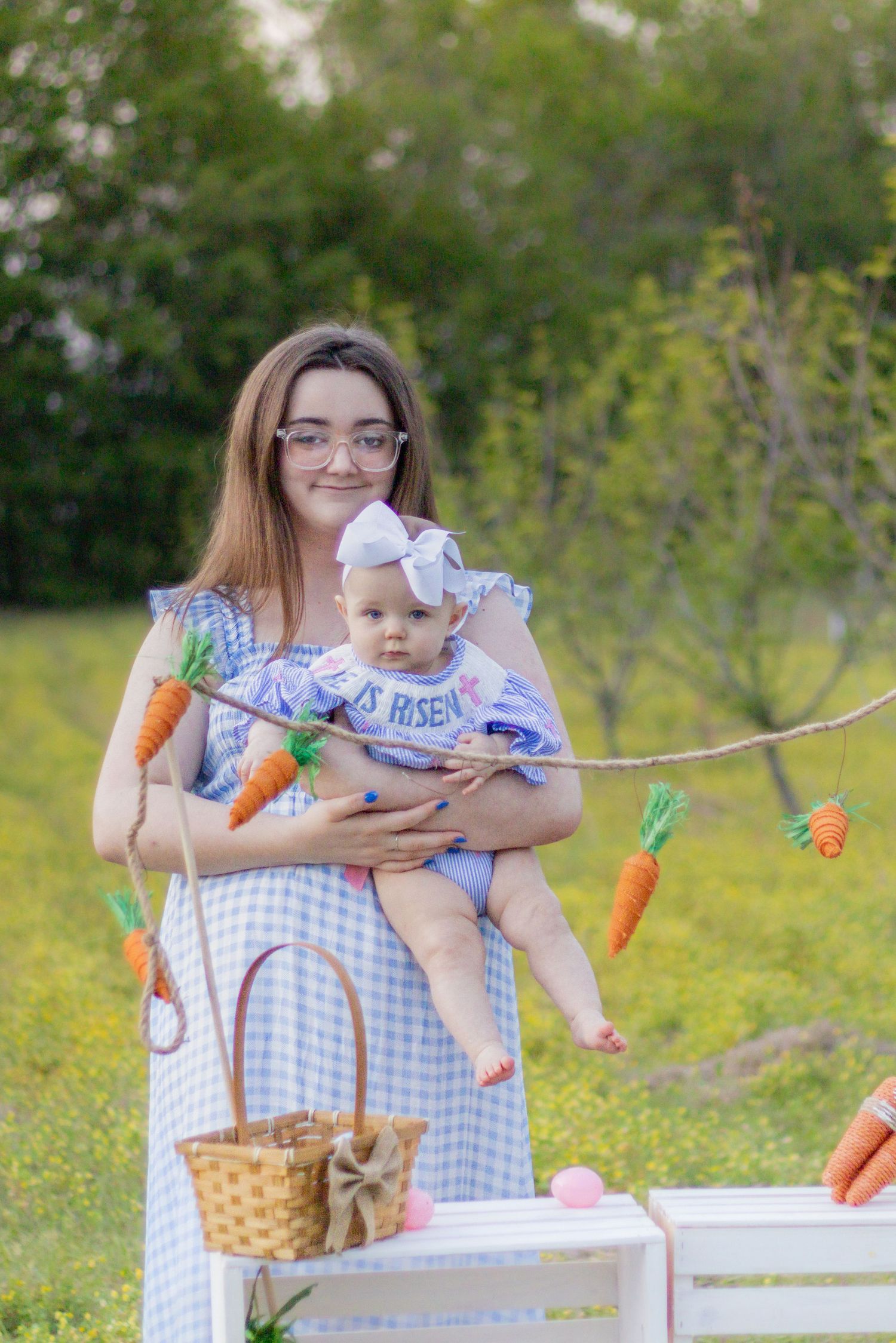Smiling mother holds baby in Easter outfit near carrot garland in yellow flower field.