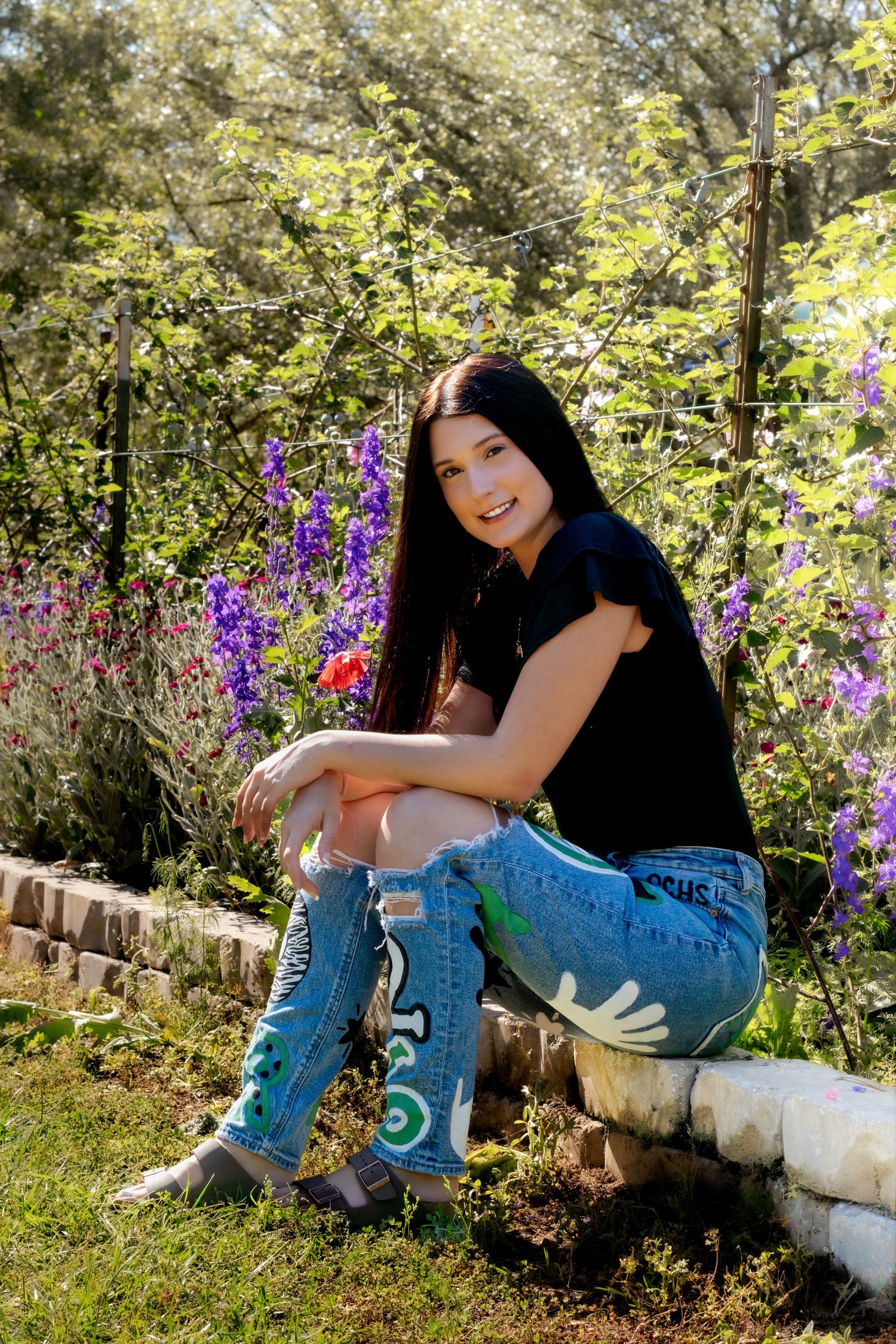 Young woman with long dark hair sits on stone border smiling, wearing black top and painted jeans, surrounded by purple flowers.