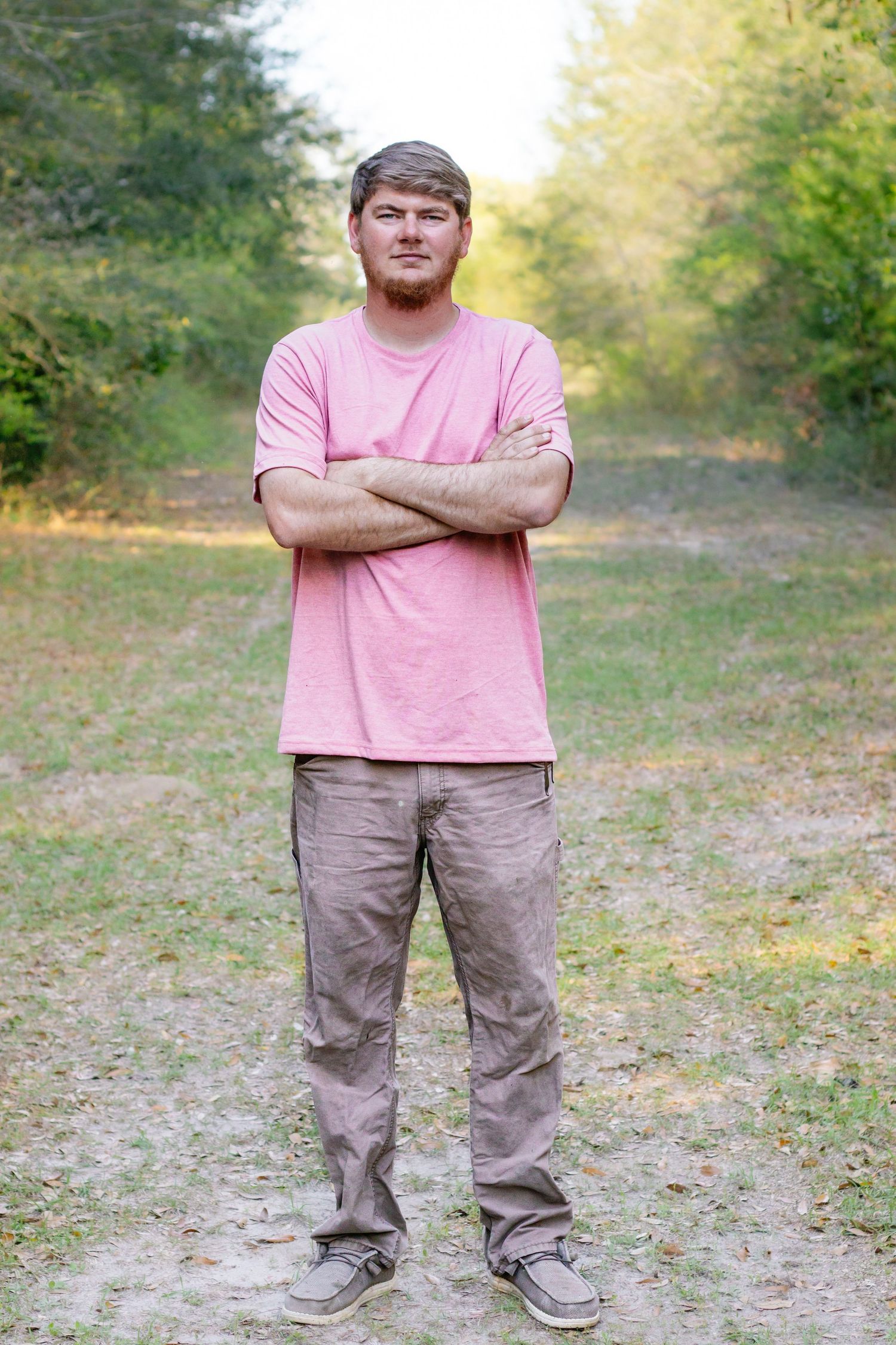 Man in pink t-shirt and khaki pants standing with arms crossed on a dirt path in a wooded area.
