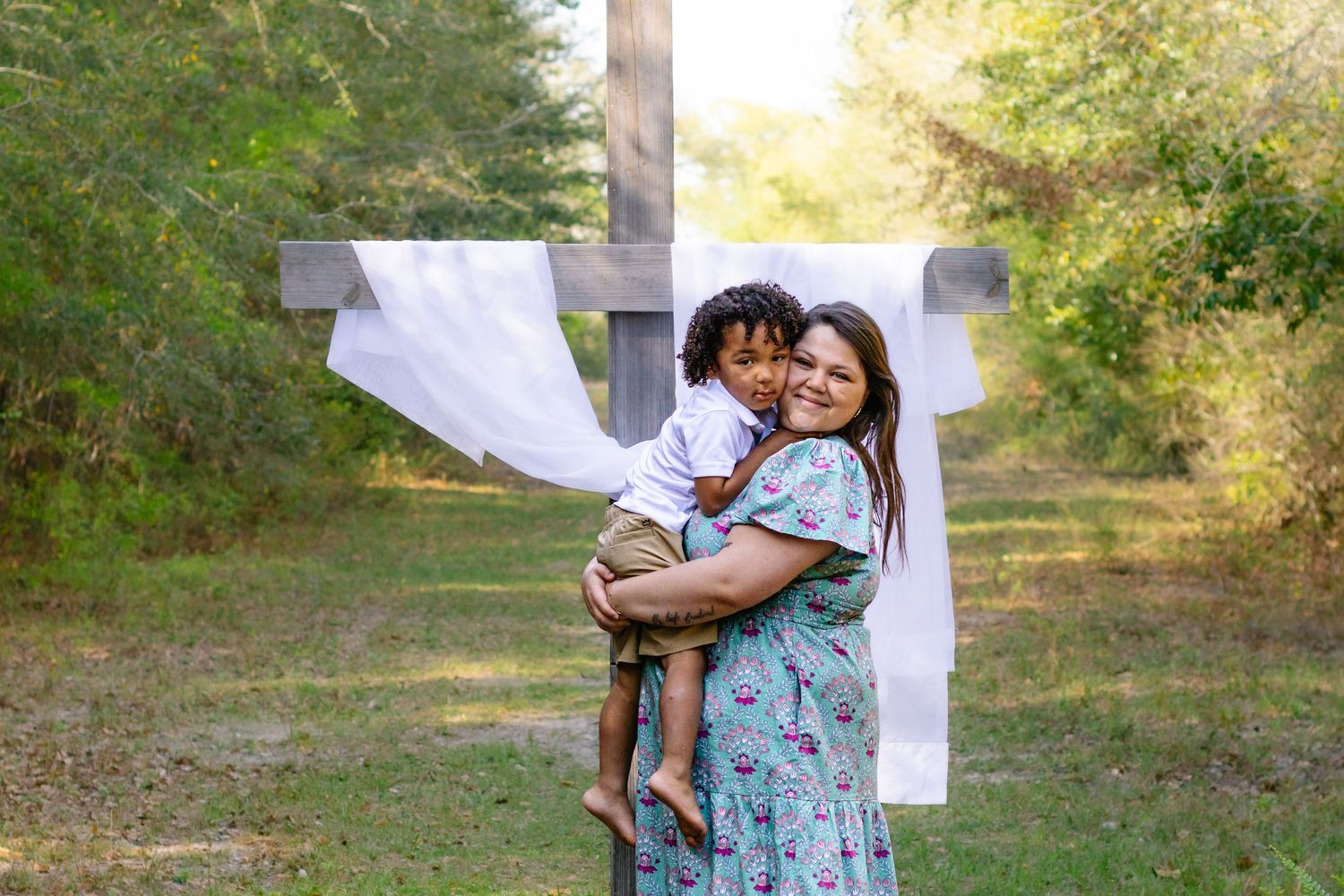 A mother holds her young child in front of a large white cross outdoors.