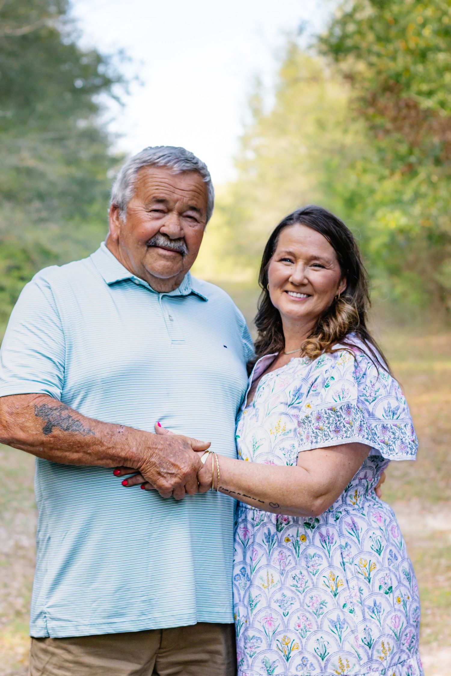 Smiling older couple holding hands outdoors with trees in the background on a sunny day.