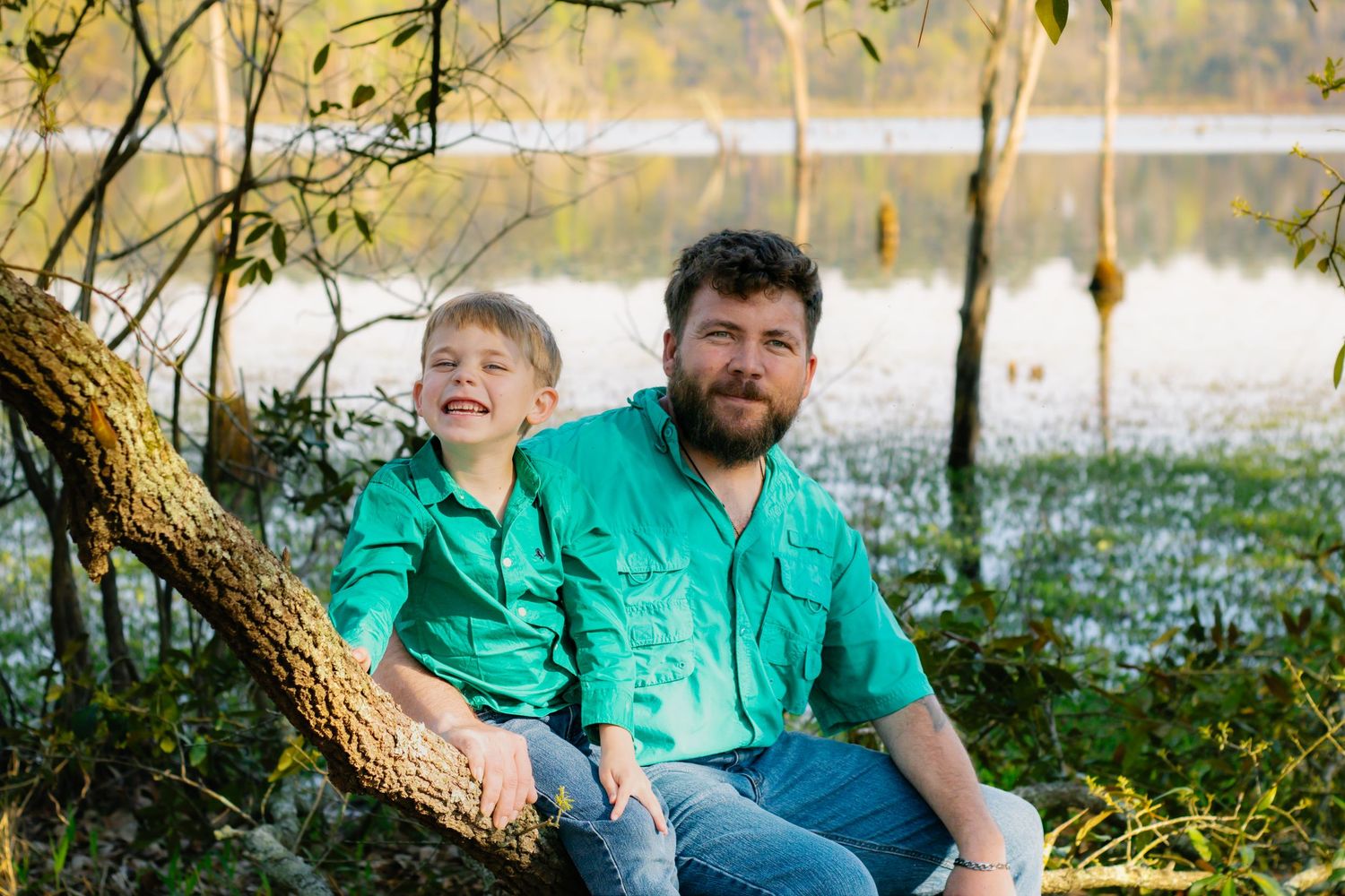 A father and young son in matching green shirts sit on a tree branch by a serene lake outdoors.
