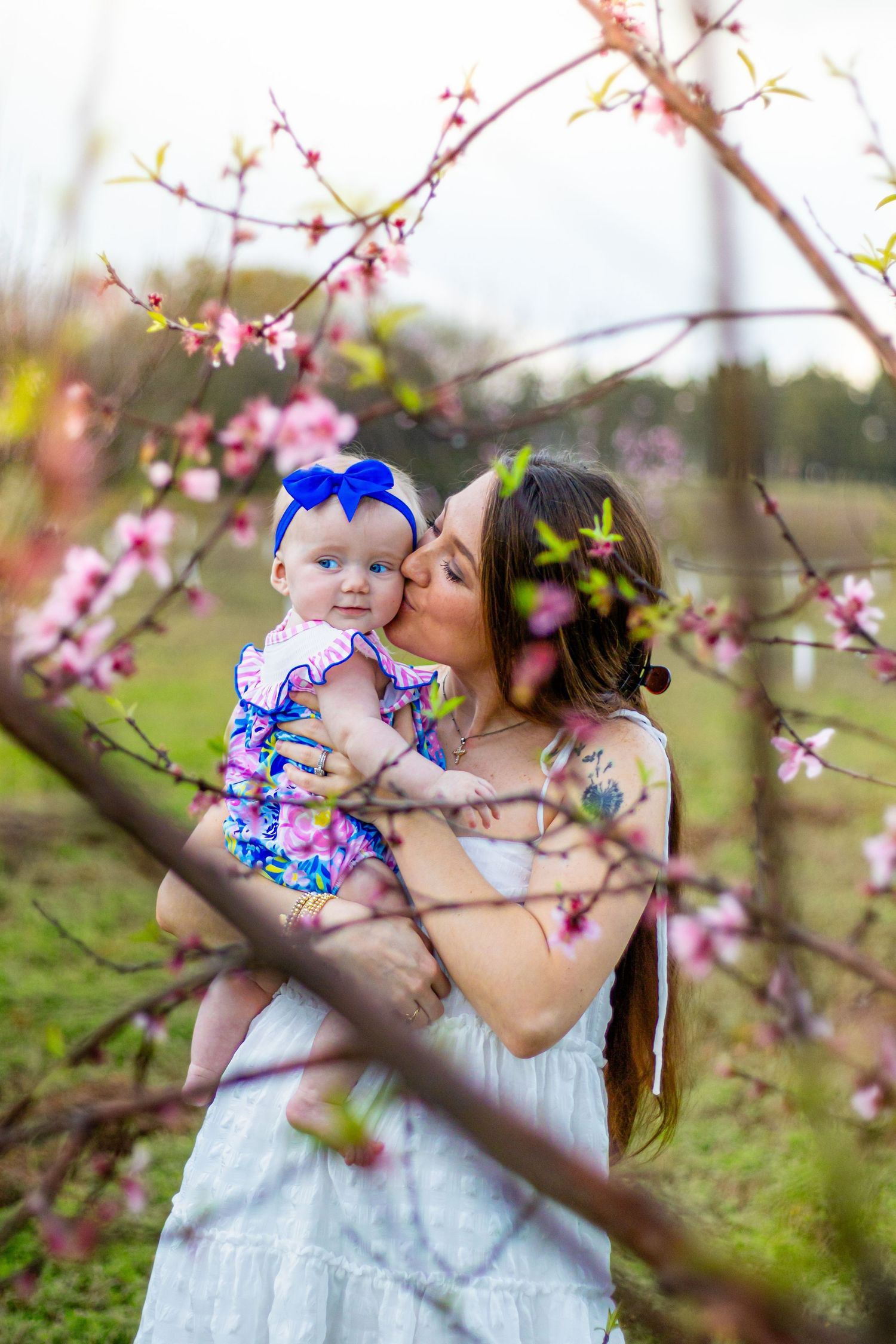 Loving mother in white dress kisses baby girl wearing blue bow headband among pink spring blossoms outdoors.