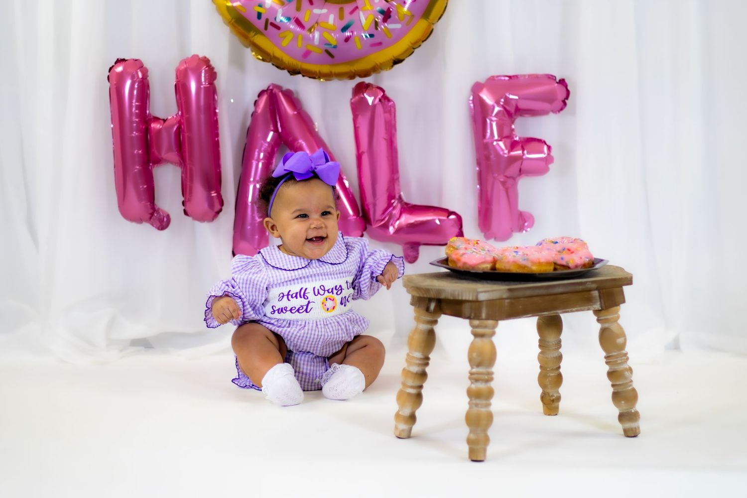 Baby girl in purple outfit sits near pink donuts on wooden stool at half birthday photo session.
