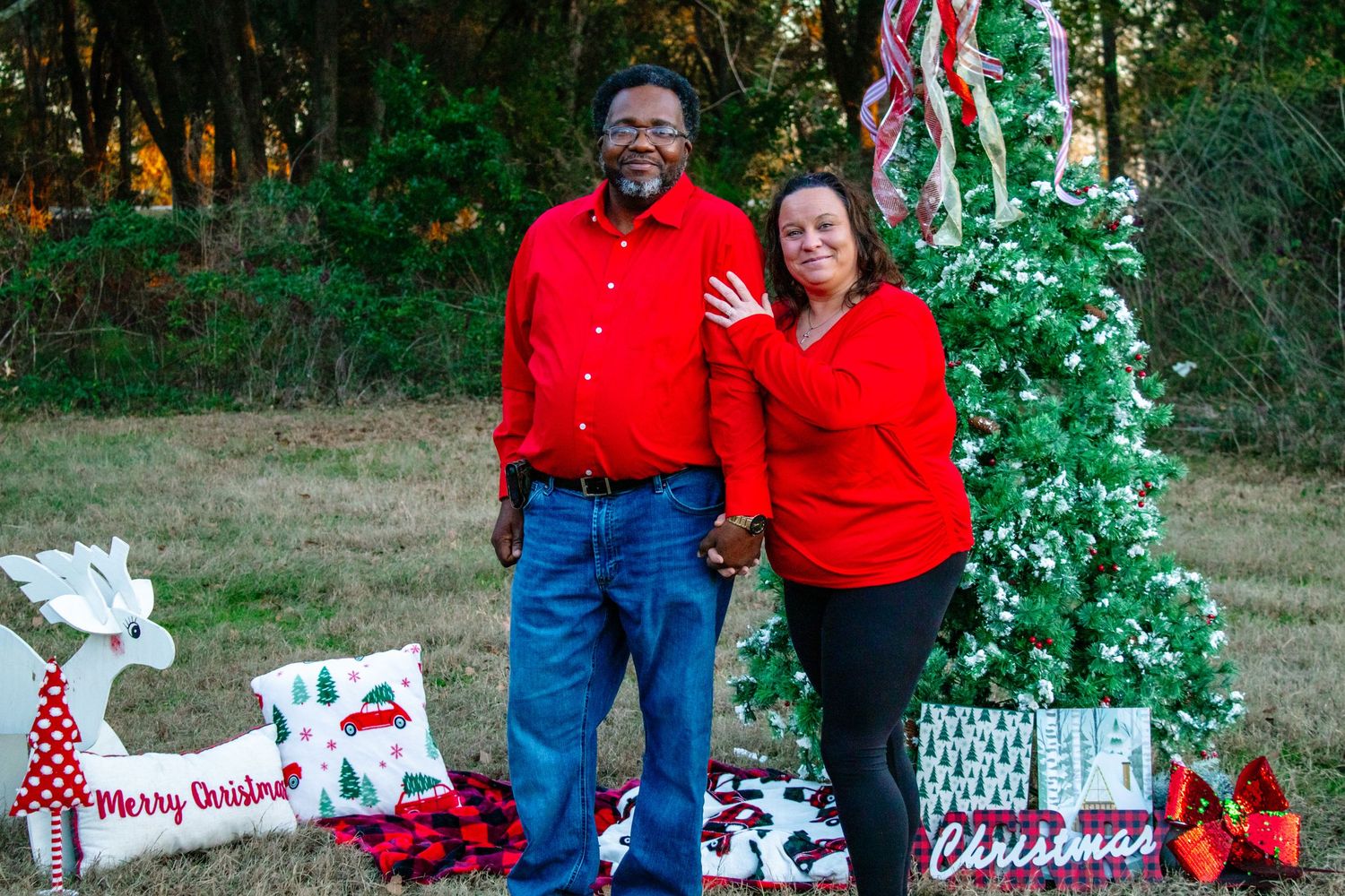 Older couple in red outfits smiling and holding hands beside a white Christmas tree with holiday decorations outdoors.