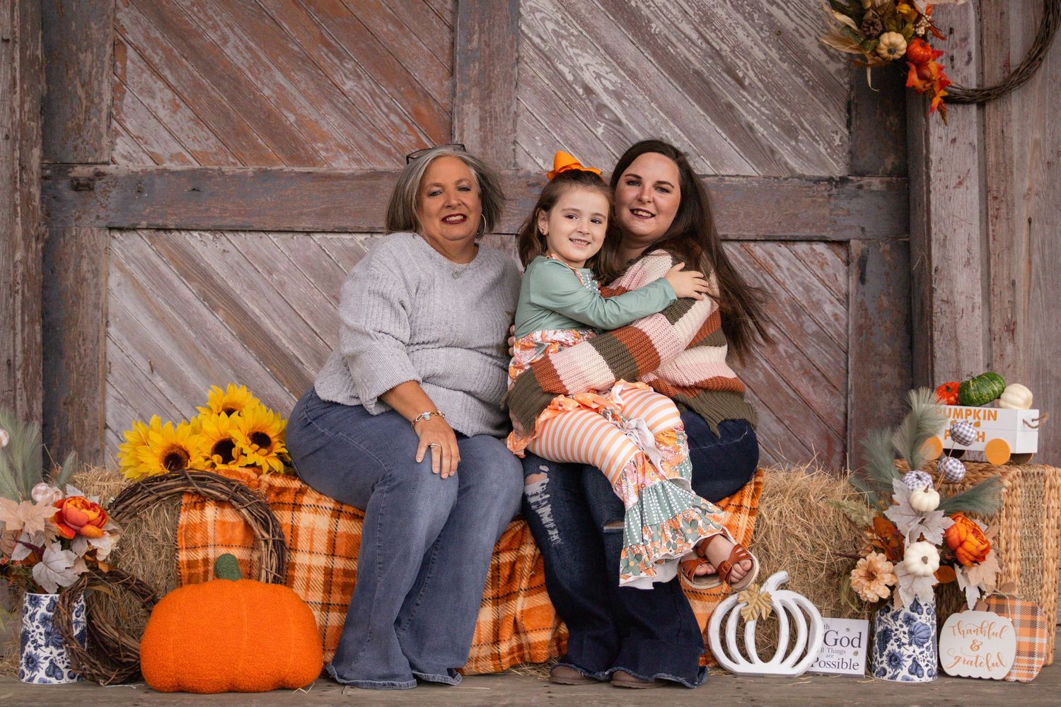 Three generations of women smile together amid fall pumpkins and sunflower decorations by rustic barn.