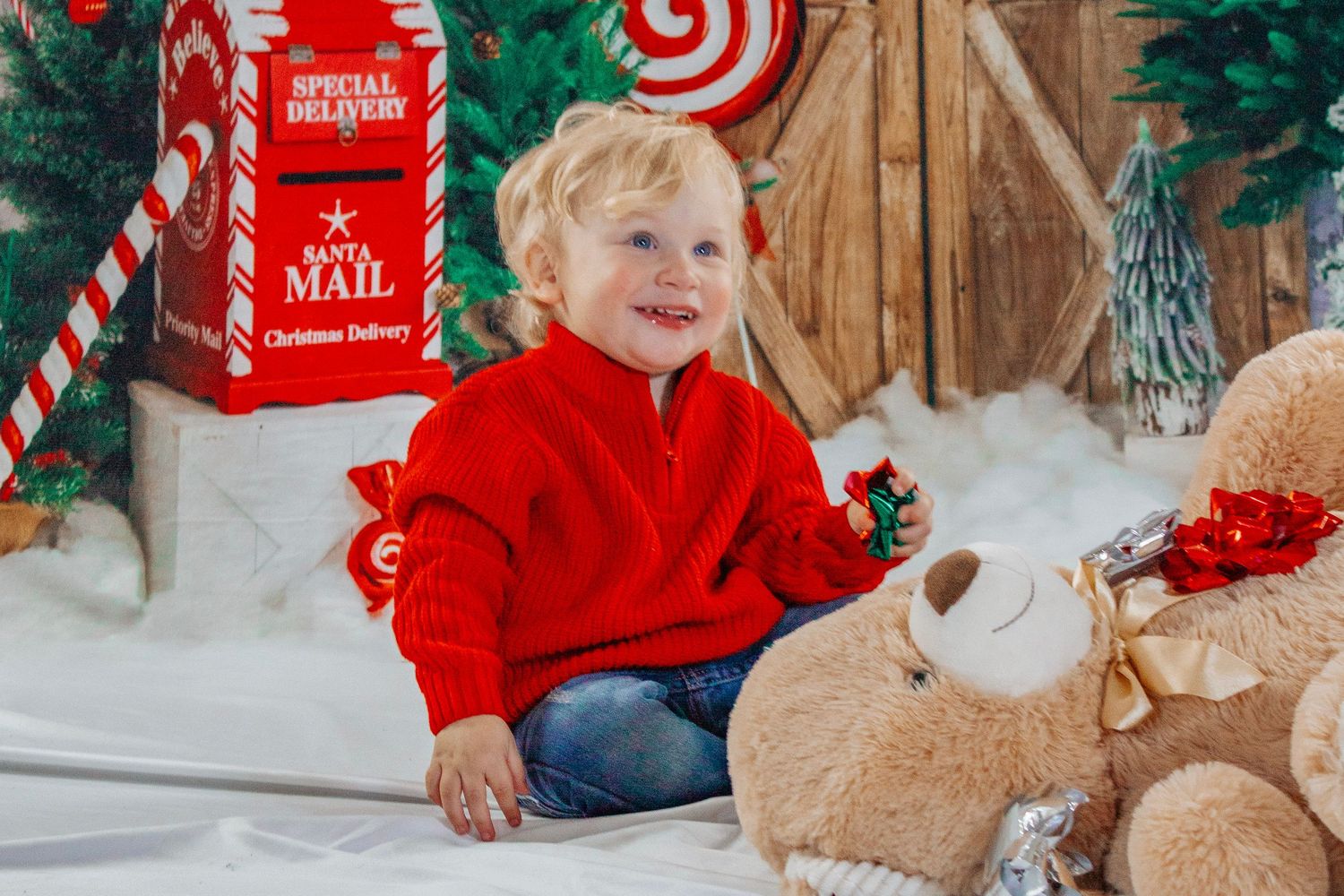 A happy toddler in a red sweater plays beside a stuffed reindeer in a festive Christmas studio setting.