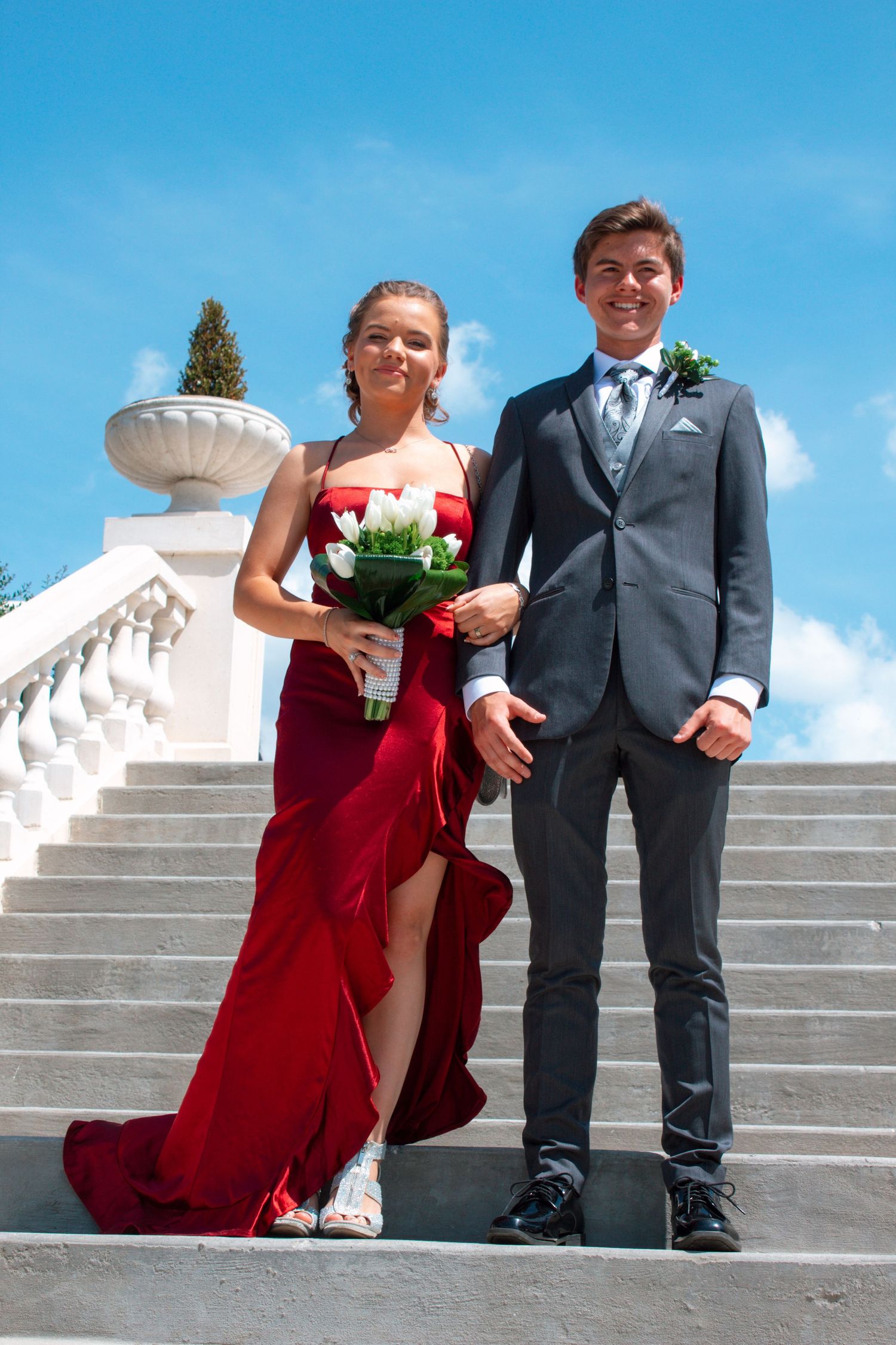 Prom couple descends white marble stairs, girl in red gown holding white roses, guy in gray suit.