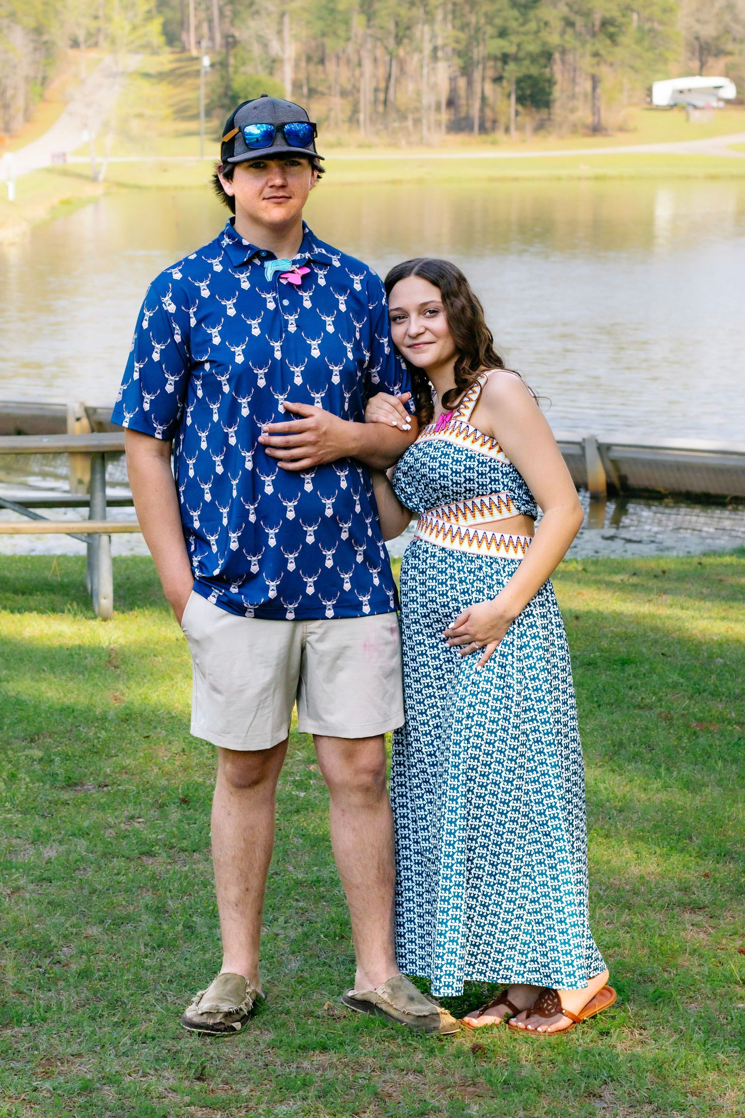 Pregnant couple poses by lake, man in blue deer shirt and khaki shorts, woman in blue patterned maxi dress.