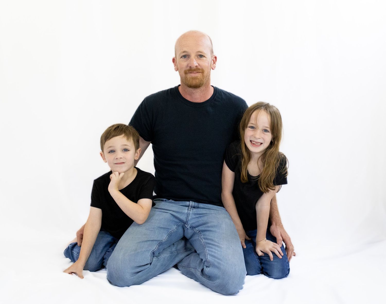 Bald man with red beard sitting on floor with two young children in matching black shirts and jeans.