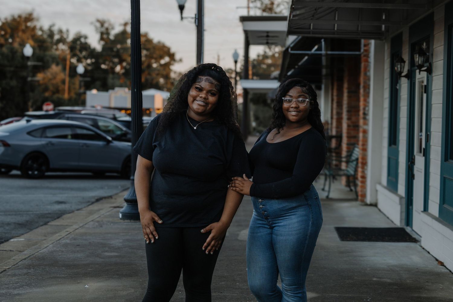 Two young Black women in black outfits smile together on a small town sidewalk.