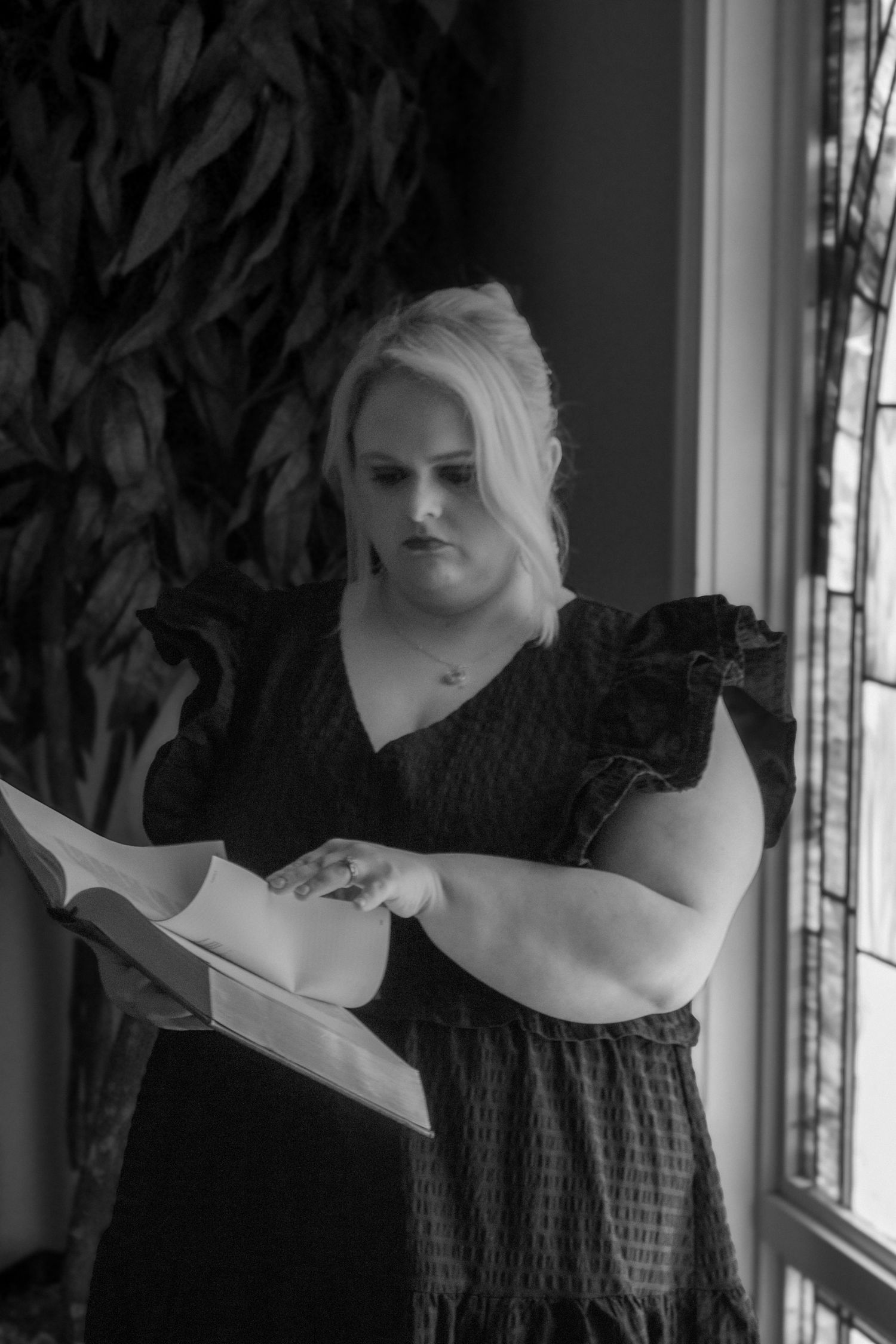 Black and white photo of blonde woman in ruffled dress reading a book near a stained glass window.