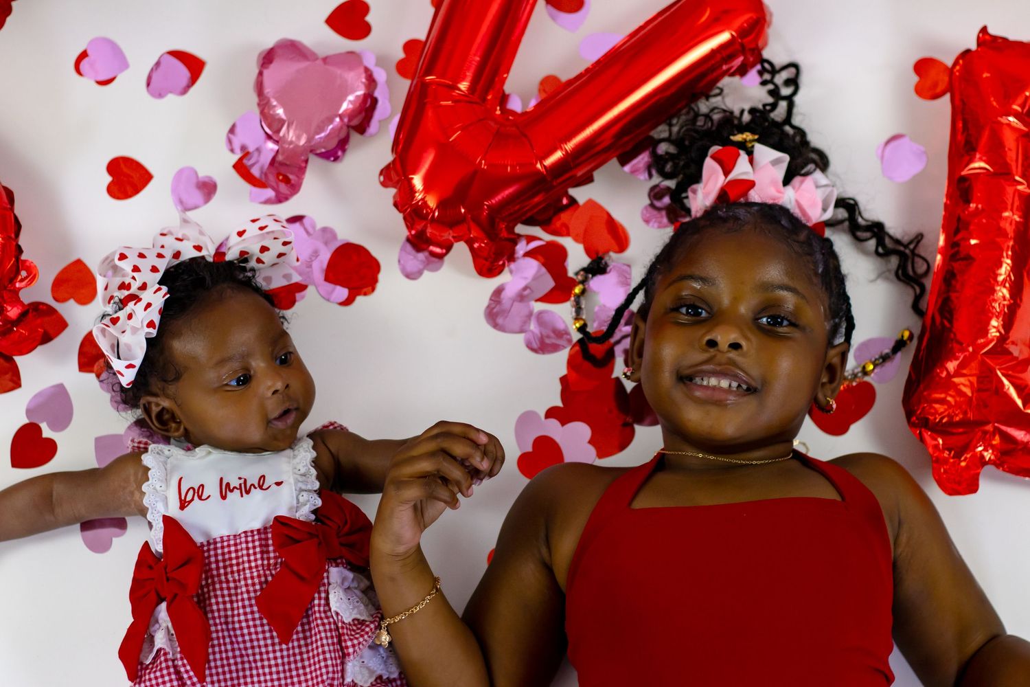Two sisters in Valentine's Day outfits laying among heart confetti with red foil balloons overhead.