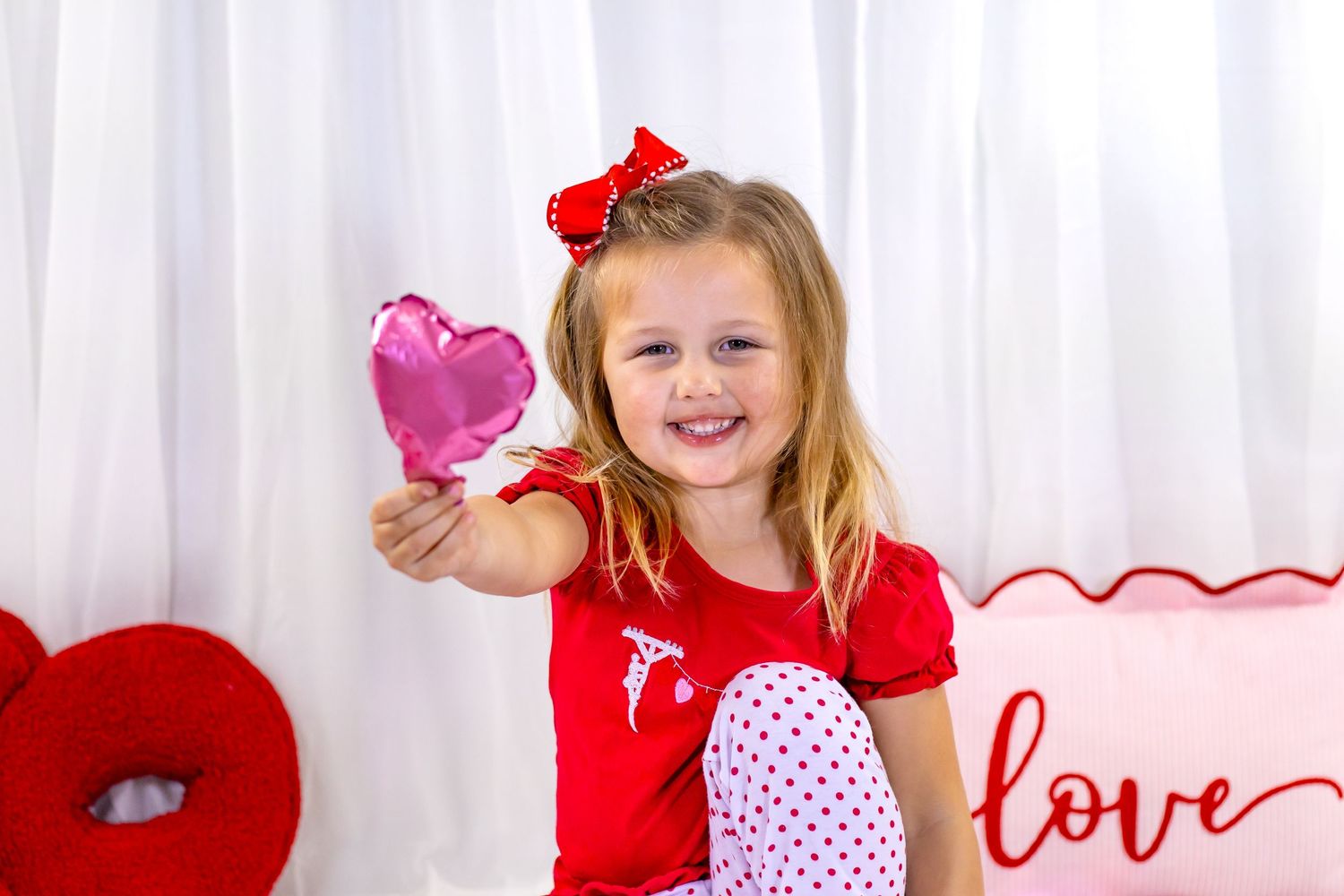 Smiling girl in red top holds a pink heart balloon near a love pillow in a Valentine's Day photo.