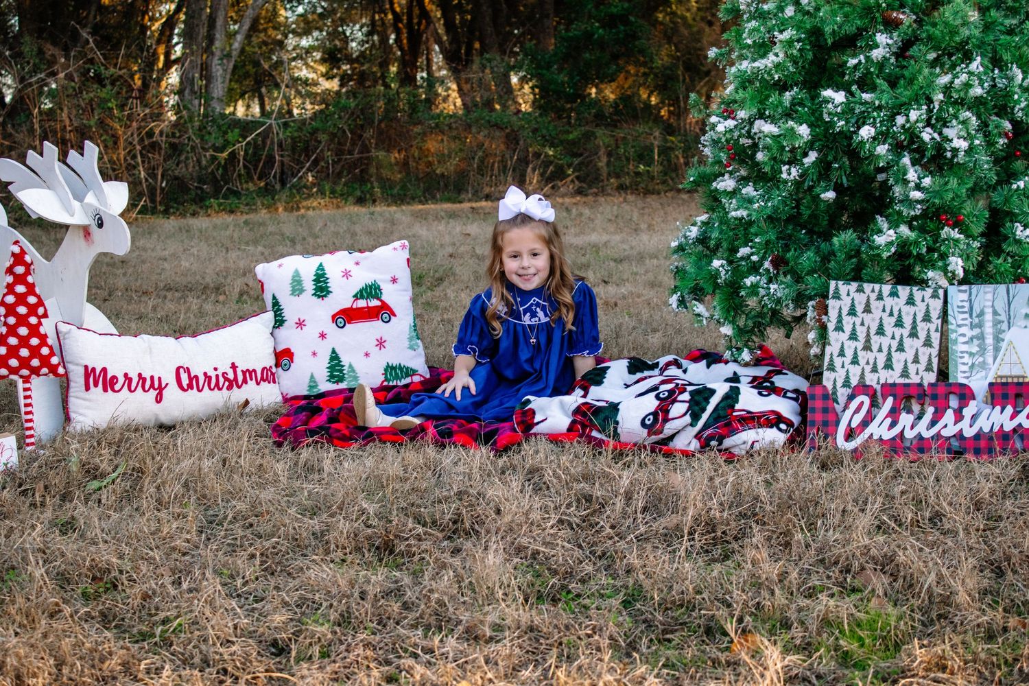 A little girl in a blue dress sits beside Christmas pillows and a decorated tree outdoors.